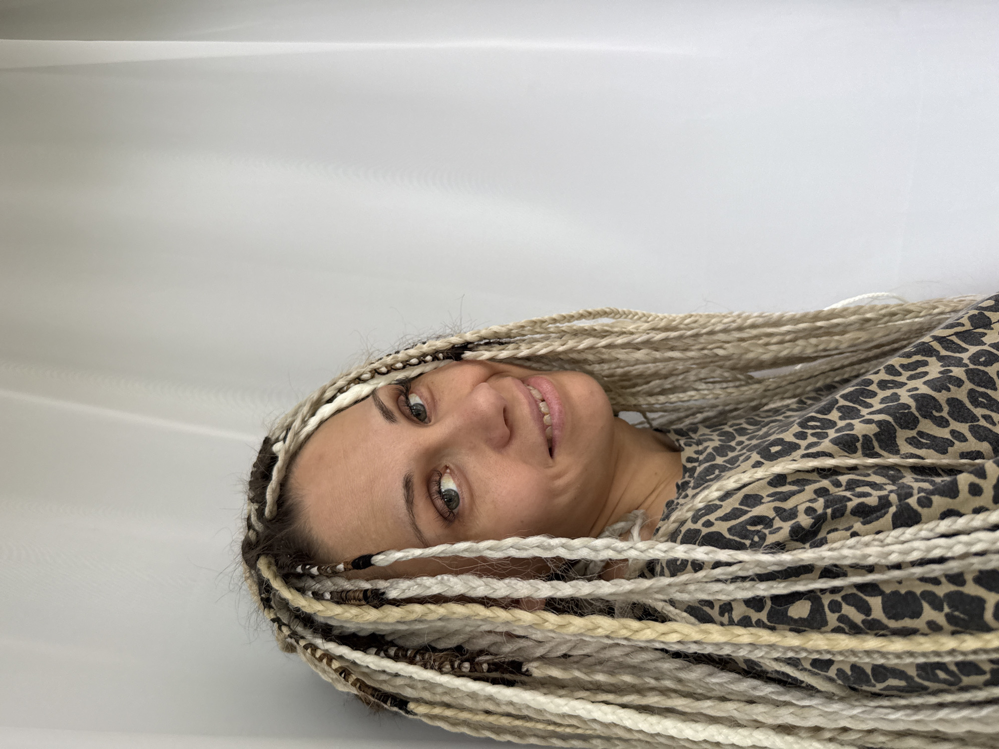 A medium shot shows a woman with blonde dreadlocks and a leopard-print shirt standing in front of a white curtain. Her head is turned to the side, and she is smiling. The background is a plain white curtain. The image appears to have been taken indoors.