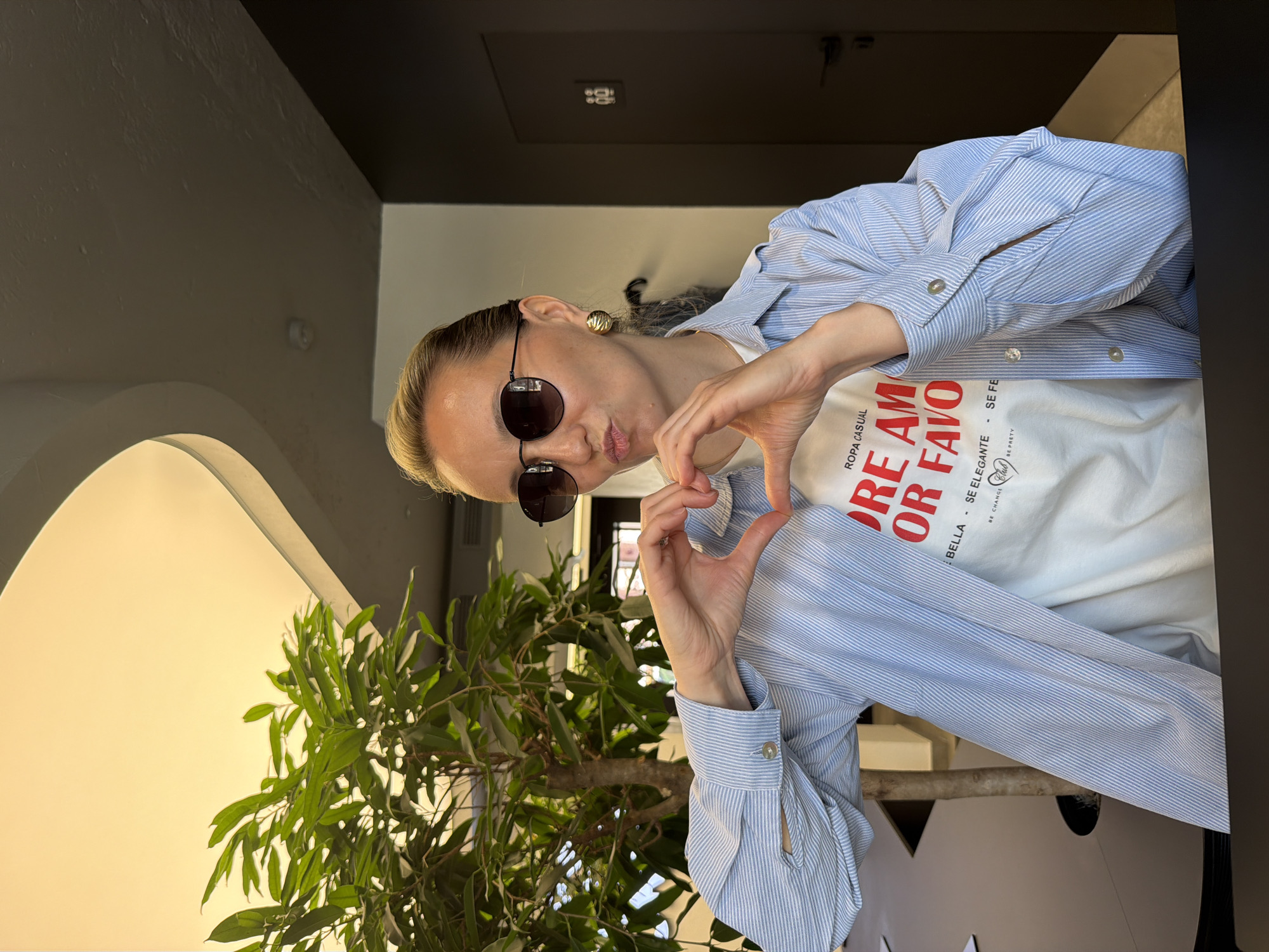 A medium shot captures a fair-skinned woman with blonde hair and sunglasses. She is making a heart shape with her hands in front of a tree. She is wearing a white t-shirt with red text and a light blue and white striped button-down shirt. The image appears to be taken outside with a white arched structure visible on the left side of the frame and a dark doorway behind the woman. The location in the city of Izhevsk, Russia, is not evident from the image.