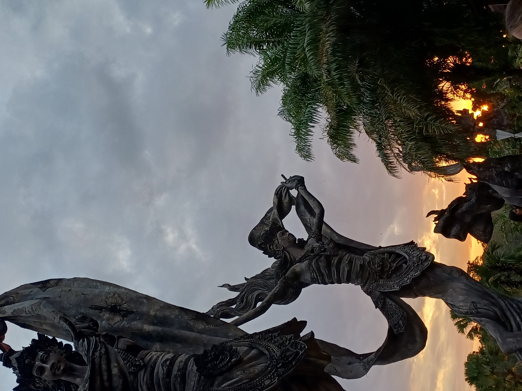 The image shows a low-angle view of statues under a sky that is gray in the upper part and light orange-yellow near the horizon, suggesting a sunset or sunrise. There are palm trees visible in the upper right of the frame. The statues appear to be Balinese in style, with intricate details on the clothing and headdresses. One statue seems to depict a male figure holding a musical instrument near his mouth. Another statue is partially visible on the left. The location is the Garuda Wisnu Kencana Cultural Park, Ubud District, Indonesia.
