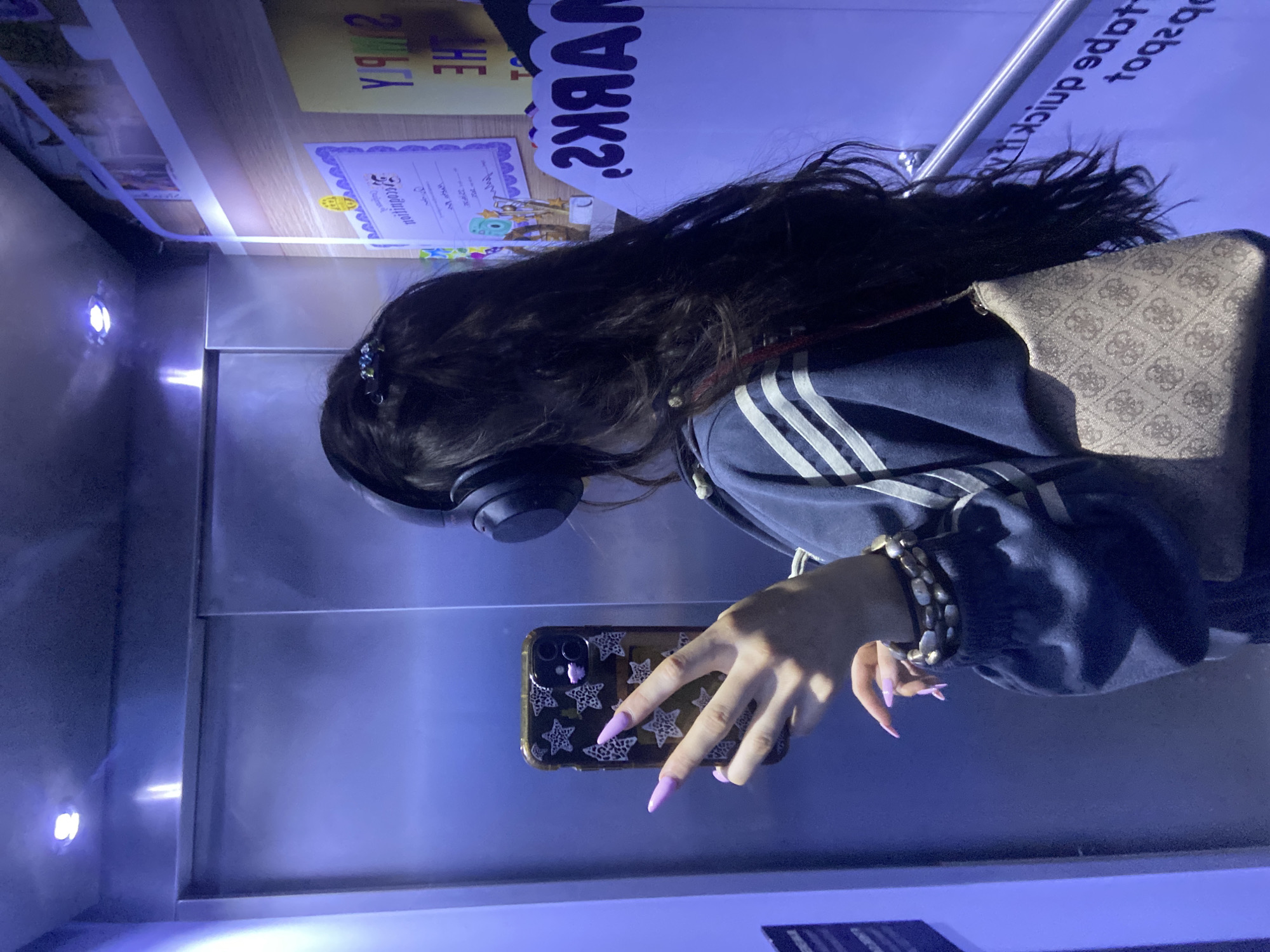 A young woman is in a commercial passenger elevator. She has long, dark, wavy hair, and is wearing over-ear headphones. She has a navy blue hooded sweatshirt with three white stripes on the sleeves, a beaded bracelet on her left wrist, and long, pointed, light-purple fingernails. She is holding a cell phone with a black case that has white star designs on it. She is taking a picture of herself with the cell phone. The elevator is made of brushed metal, and there are small, round lights in the ceiling. There are two posters attached to the elevator's back wall. The background of the images are blurry, and therefore unidentifiable.
