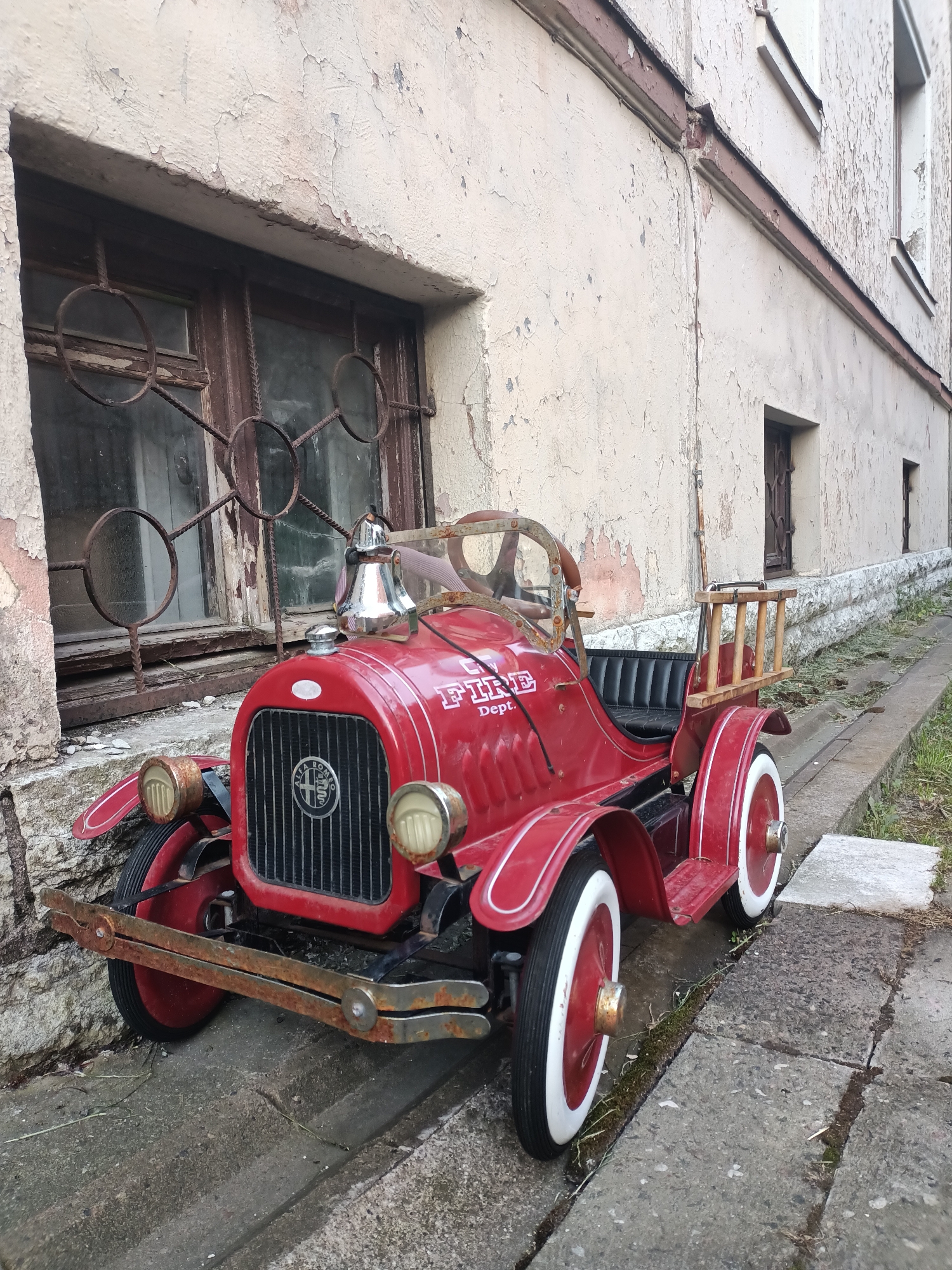 A bright red toy fire truck is parked next to a weathered, pale beige building. The building has large, dark-paned windows covered with ornate, rusty metal grates. The paint on the building is peeling and cracked, revealing older layers of paint and brickwork underneath. A concrete curb runs along the base of the building, with patches of grass and weeds growing in the cracks. The toy truck has a prominent Alfa Romeo badge on its grille and the word "FIRE" with "Dept." below it in white lettering on its side. It features a chrome bell on top, a wooden steering wheel, black leather-look seating, and a small wooden ladder rack at the back. The truck's wheels are red with white rims. The scene appears to be outdoors, possibly in an alley or courtyard. The overall impression is one of age and disuse, with the vibrant toy truck contrasting with the dilapidated surroundings. The location is Tallinn, Estonia.