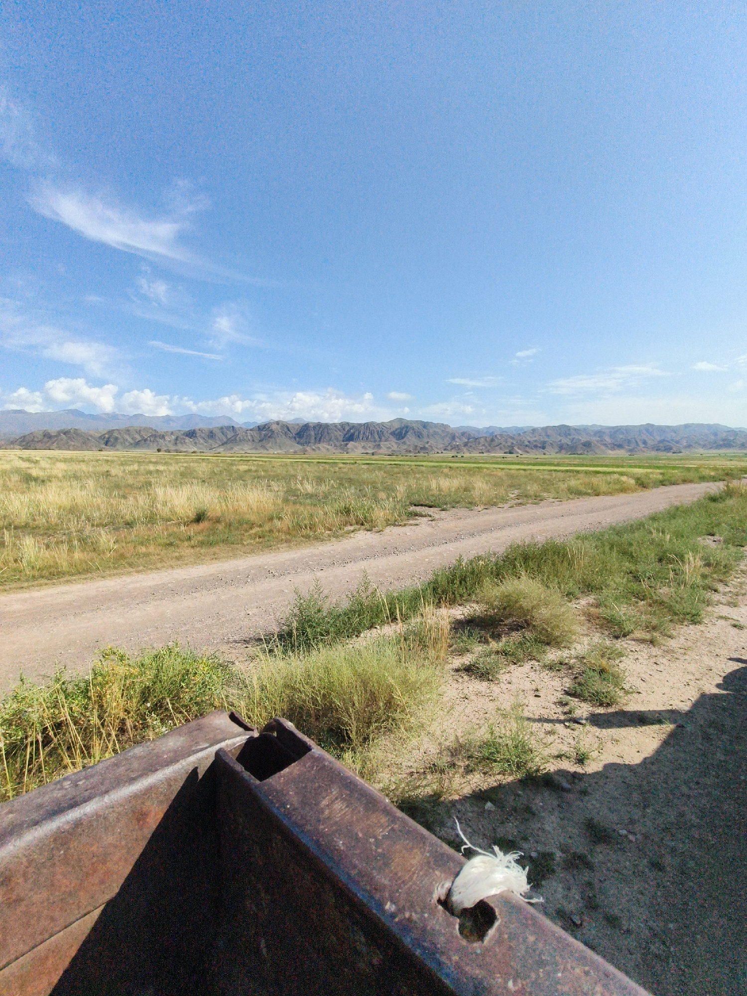 The image depicts a wide, arid landscape under a clear blue sky with sparse clouds. In the foreground, the rusted metal structure of what appears to be a truck bed or trailer is visible, with a piece of white twine protruding from a hole in the metal. A dirt road cuts through the middle ground, leading towards a distant mountain range. The surrounding terrain is a mix of dry grass and sparse vegetation. The overall impression is one of a remote, undeveloped area.