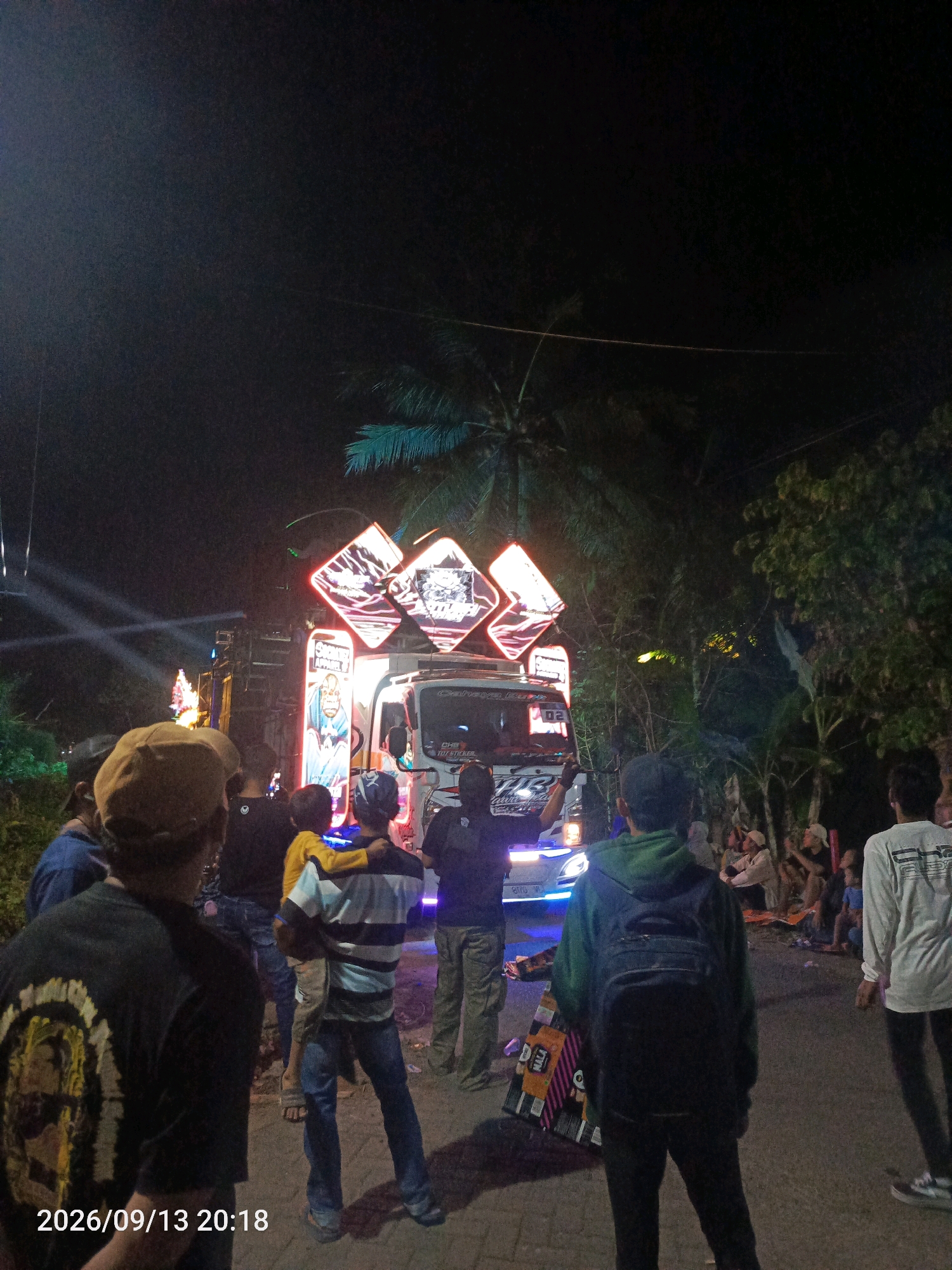 At night in Kebondalem, Indonesia, a group of people are gathered on a street next to a modified truck adorned with large, glowing diamond-shaped signs and colorful graphics. The truck has a prominent sound system and is lit with bright neon lights, suggesting it is part of a parade or event. Some individuals are standing and watching the truck, while others appear to be socializing. A man in the foreground wears a black t-shirt with a graphic design and a tan baseball cap. A child is being held by an adult wearing a striped shirt. The environment is dimly lit, with trees and vegetation visible in the background, indicating an outdoor setting. The timestamp on the image reads 2026/09/13 20:18.