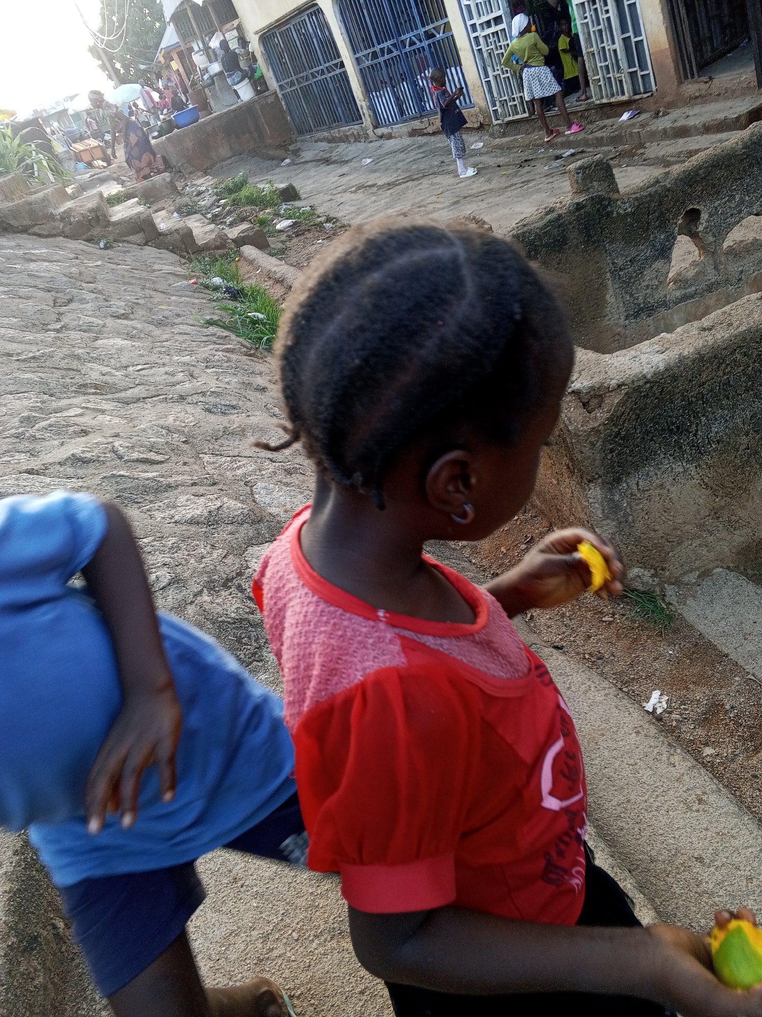 The image shows a partially visible child in a blue shirt and shorts and a child in a red shirt holding a yellow fruit. The background shows a street in Gwong, Nigeria. There are shops with metal shutters and people are walking and carrying objects. The street is made of stone and cement, and there is a small amount of vegetation visible.