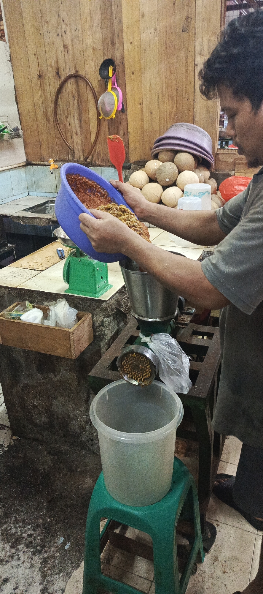 A man is pouring ingredients from a blue bowl into a metal grinder in Sukaresmi, Indonesia. The grinder is attached to a metal stand with a green base. A clear plastic bucket sits below the grinder on a green stool, collecting the processed ingredients. The background features a wooden wall with various items hanging on it, including strainers and a red spatula. Piles of light-colored, round objects, possibly coconuts or potatoes, are visible behind the grinder. The scene appears to be set in a market or food preparation area.