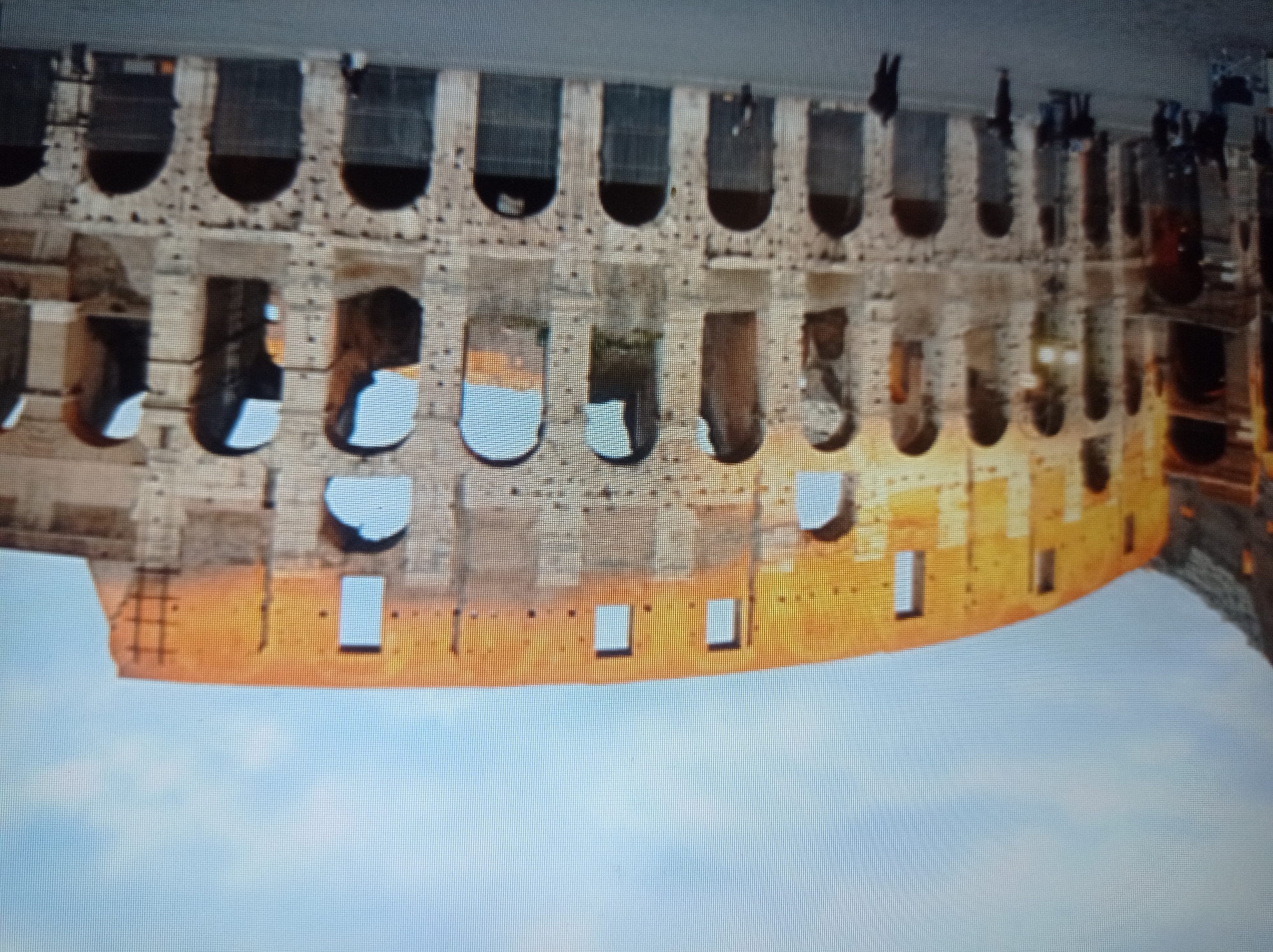 The image shows a wide-angle view of the Colosseum in Rome, Italy. The ancient Roman amphitheater is predominantly made of stone and brick, with its iconic arched structure visible. Sunlight illuminates the upper portions of the structure, casting a warm glow on the weathered stone. In the foreground, a paved area shows a few scattered individuals, some appearing to be tourists, with no discernible interactions. The sky above is partially cloudy, with a pale blue hue. The location is Rome, Italy, not Mukachevo, Ukraine.