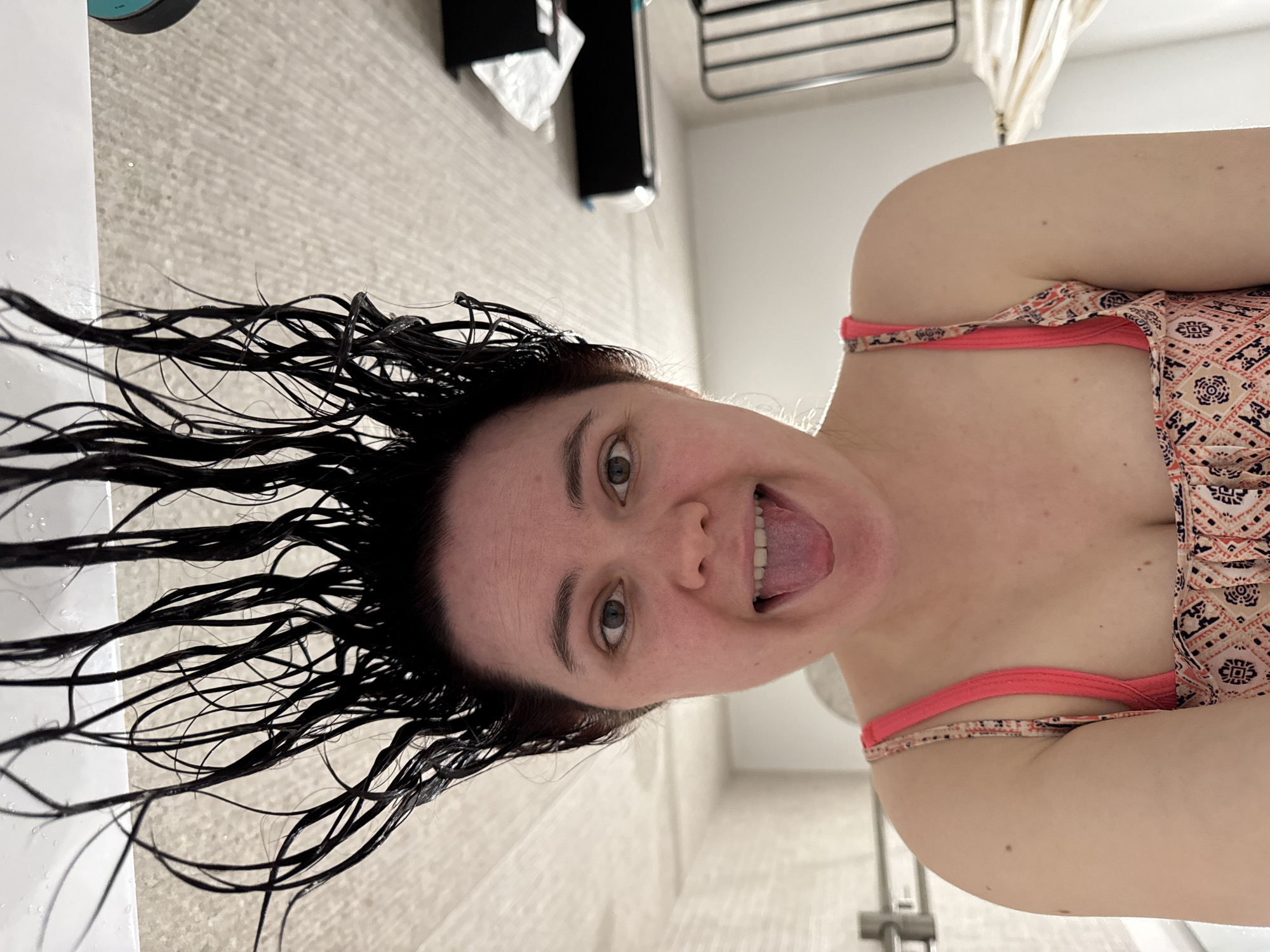 A selfie photograph shows a woman with wet, dark hair hanging down towards the left side of the frame. She has fair skin, and her eyes are open and looking at the camera. Her mouth is open and her tongue is sticking out. She is wearing a swimsuit with a peach and black pattern, and a pink strap is visible. The background includes a white wall and a shower. Part of a vanity, tissue box, and towel rack are also visible. The location is not specified and thus cannot be inferred as being in the city of Izhevsk, Russia.