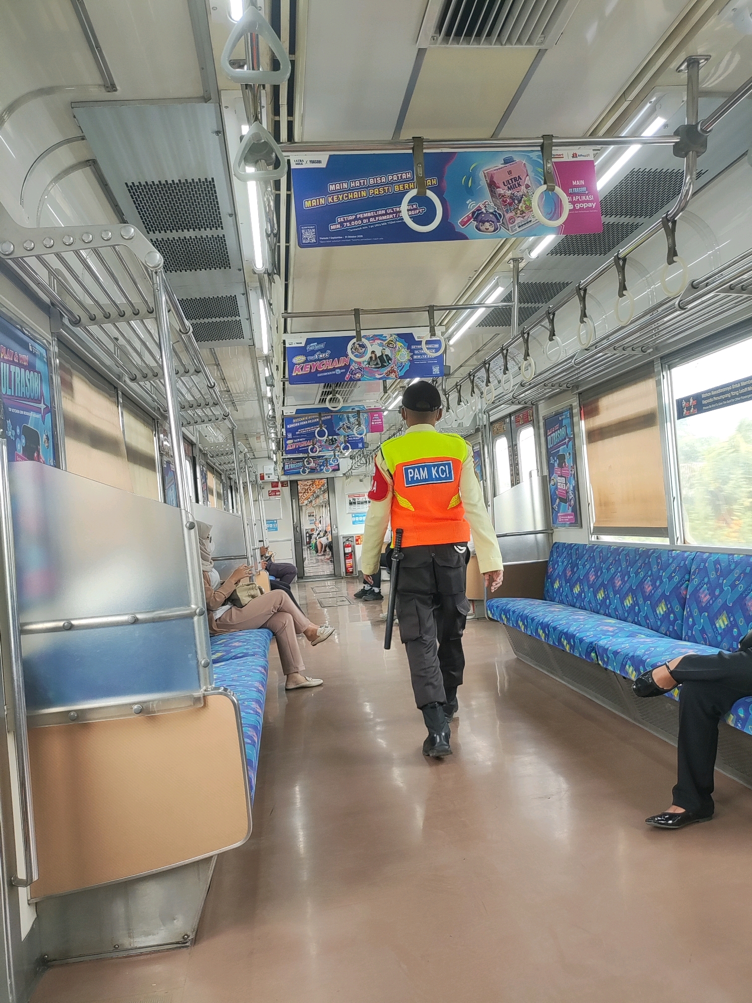 The image depicts the interior of a commuter train carriage, likely on the KCI Commuter Line. The scene is captured from a slightly elevated perspective, looking down the aisle of the train. The lighting is bright, indicating fluorescent lighting overhead.
In the foreground, a uniformed individual, identified by a reflective orange vest with "PAM KCI" clearly printed on the back in blue lettering, walks away from the camera. They are wearing dark trousers, black boots, and a black cap. A dark baton is attached to their belt on their left side.
Seated on the left side of the carriage are several passengers. One woman, wearing a light-colored hijab and matching pantsuit, is looking at her phone. Another individual is also seated further down the bench. The seats are upholstered in a blue fabric with a patterned design. The carriage walls are metallic and feature advertisements.
On the right side of the carriage, a blue upholstered bench seat with a similar pattern is visible. A person in black trousers and black shoes is seated on this bench, their legs crossed.
The floor of the carriage is a light brown, polished surface that reflects the overhead lights. Overhead, metal handrails and luggage racks are prominently displayed. Numerous advertisements are affixed to the ceiling and walls of the carriage, featuring promotional content for products and services, including one for "ULTRA MILK."
The environment suggests a public transportation setting in Indonesia, consistent with the KCI Commuter Line operating in the Greater Jakarta area, which includes cities like Tambun. The overall impression is that of a typical journey on Indonesian public transport.