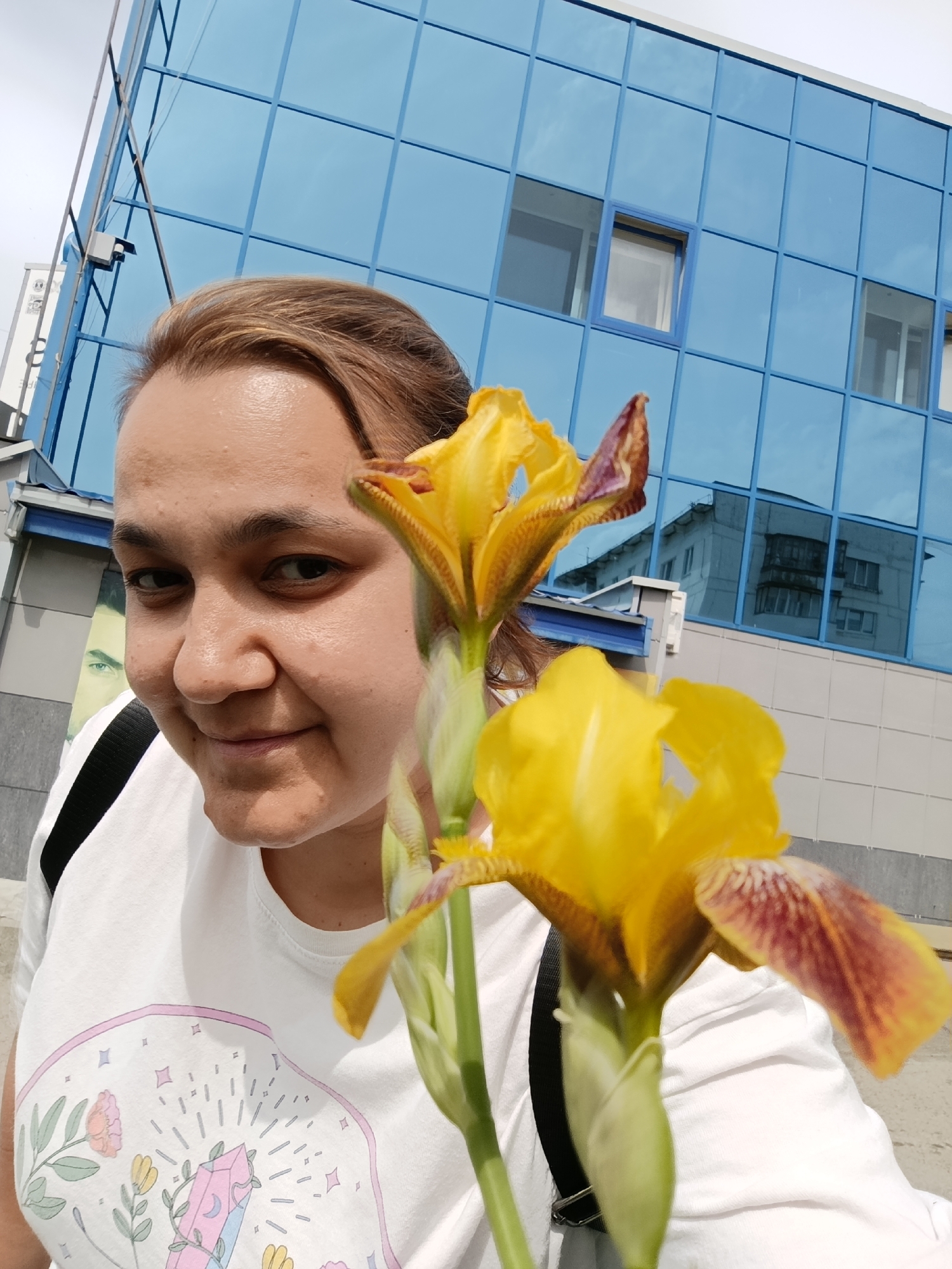 A woman is holding two yellow irises in front of a blue building. The building has a grid of windows, some of which reflect another building. The woman is fair-skinned with brown hair, wearing a white t-shirt with a printed design. She has a black strap over her shoulder. In the background, there is part of a mural on a building. This scene takes place in Chaikovsky, Russia.