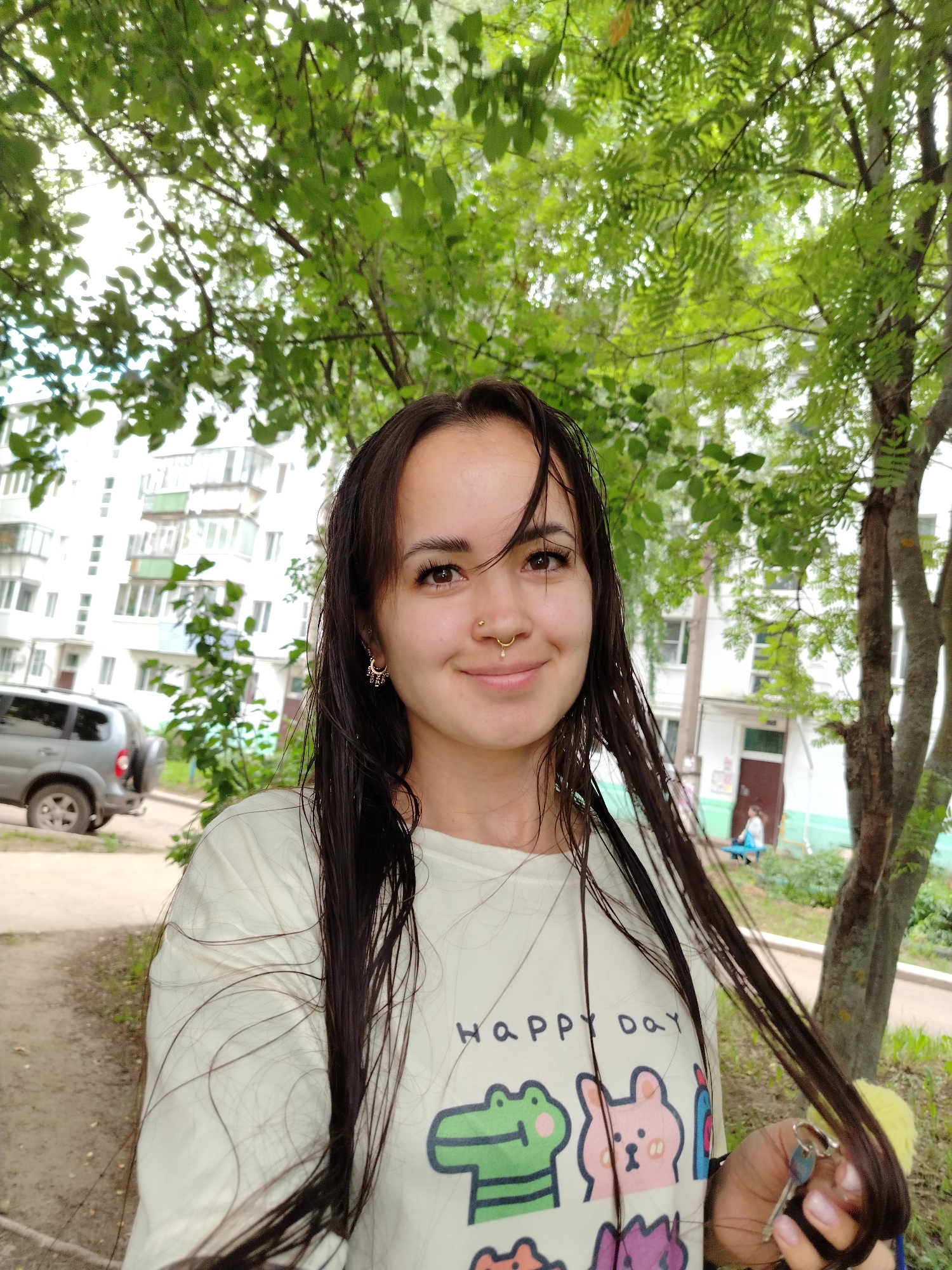 The image shows a young woman with long, dark, wet hair, wearing a light-colored t-shirt with cartoon animals printed on it, standing outdoors. She is smiling and holding a set of keys in her right hand, which is partially obscured by her hair. Her hair appears to be damp, suggesting she may have recently washed it or been in the rain.
In the background, there is a multi-story residential building with balconies. To the left of the woman, a silver SUV is parked. The scene is set against a backdrop of lush green trees and foliage, indicating an outdoor urban environment. The sky is overcast.
The location is identified as the city of Chaikovsky, Russia. The residential building in the background is typical of Soviet-era architecture commonly found in Russian cities. The presence of the SUV and the general urban setting further confirm this location. The overall atmosphere is casual and relaxed, with the woman appearing to be in a public outdoor space.