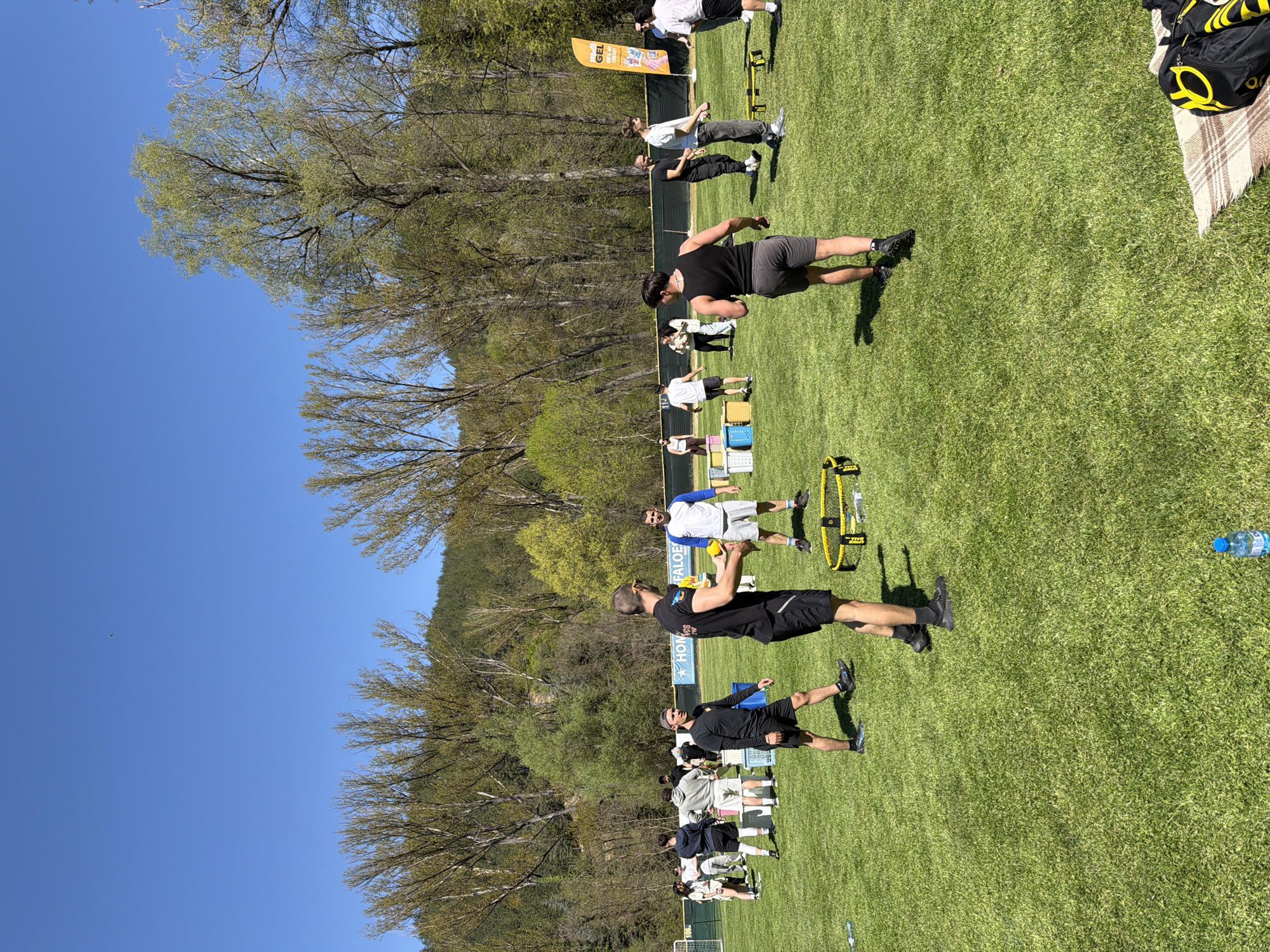 The scene depicts a group of people engaging in physical activity on a grassy field, likely a training session or workout. The location appears to be outdoors, with trees and a mountain visible in the background.
**People:** The group consists of several individuals, mostly men, dressed in athletic wear. Some are standing, others are moving, and a few seem to be using training equipment.
**Subjects:** The primary subject is the group's activity, which seems to be a form of athletic training or exercise.
**Objects:** Objects present include training equipment such as agility ladders and hurdles, water bottles, bags, and a banner.
**Actions:** People are seen performing exercises, possibly agility drills or warm-ups. Some are interacting with the equipment, while others seem to be observing or instructing.
**Interactions:** There are interactions between the people, with some likely instructing or assisting others. Interactions with training equipment is apparent.
**Environment:** The environment is an open, grassy field with trees and a mountain in the background. It appears to be a sunny day with clear blue sky. The location is identified as Blagoevgrad, Bulgaria, based on the content.