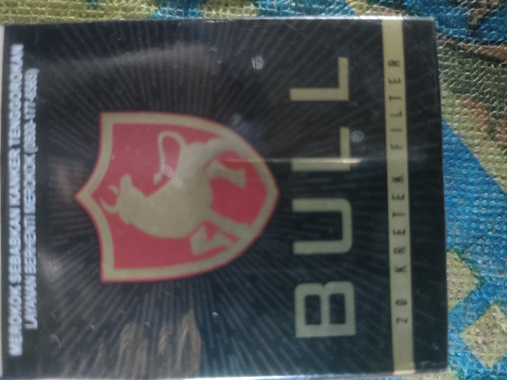 The image shows a close-up view of a pack of "Bull" brand kretek cigarettes. The pack is primarily black with a stylized red and beige bull's head emblem on the left side. The text "MEROKOK SEBABKAN KANKER TENGGOROKAN LAYANAN BERHENTI MEROKOK (0800-177-6565)" is printed vertically in white on the left side, indicating health warnings in Indonesian. Large, bold, light yellow letters spelling "BULL" are prominently displayed vertically on the right side of the pack. A vertical yellow stripe on the far right contains the text "20 KRET EK FILTER" printed vertically. The background consists of a textured surface, likely a woven or fabric material, in shades of blue, yellow, and green. There are no people or discernible city landmarks visible. The lighting suggests an indoor environment with artificial light sources. The image does not provide any specific geographical information or context that would place it in the city of Padang Dalom, Indonesia.