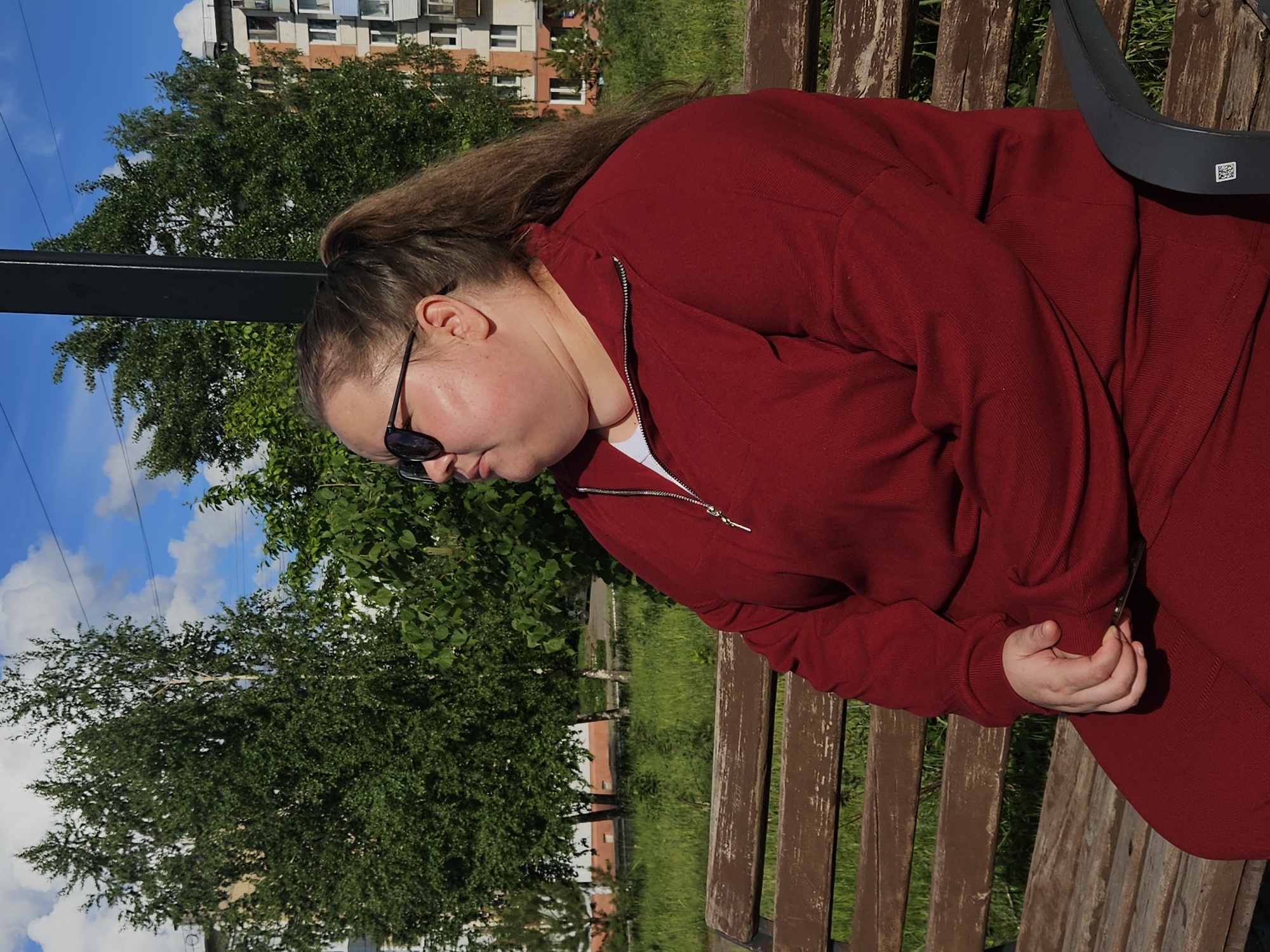 A person is sitting on a wooden bench outdoors. The person is wearing a dark red sweatshirt and matching pants. The person has sunglasses on and their hair is pulled back. Behind the person are green trees, a blue sky with white clouds, and a building. The photo was taken in the city of Chaikovsky, Russia.