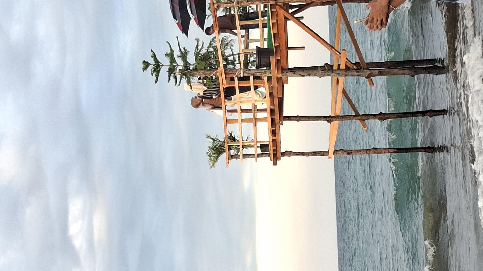 This image shows a wooden pier extending over the ocean at a beach in Lhokseumawe, Indonesia. The pier has a railing and is supported by wooden posts. On the pier, there are three people. Two individuals, appearing to be women wearing hijabs, are standing together and looking out at the water. A third person, a man, is also on the pier. Several potted plants and two umbrellas are also visible on the pier. The ocean has waves and appears somewhat rough. The sky is overcast with clouds. A person's hands holding a phone are visible in the lower right corner of the frame, suggesting they may be taking the photograph.