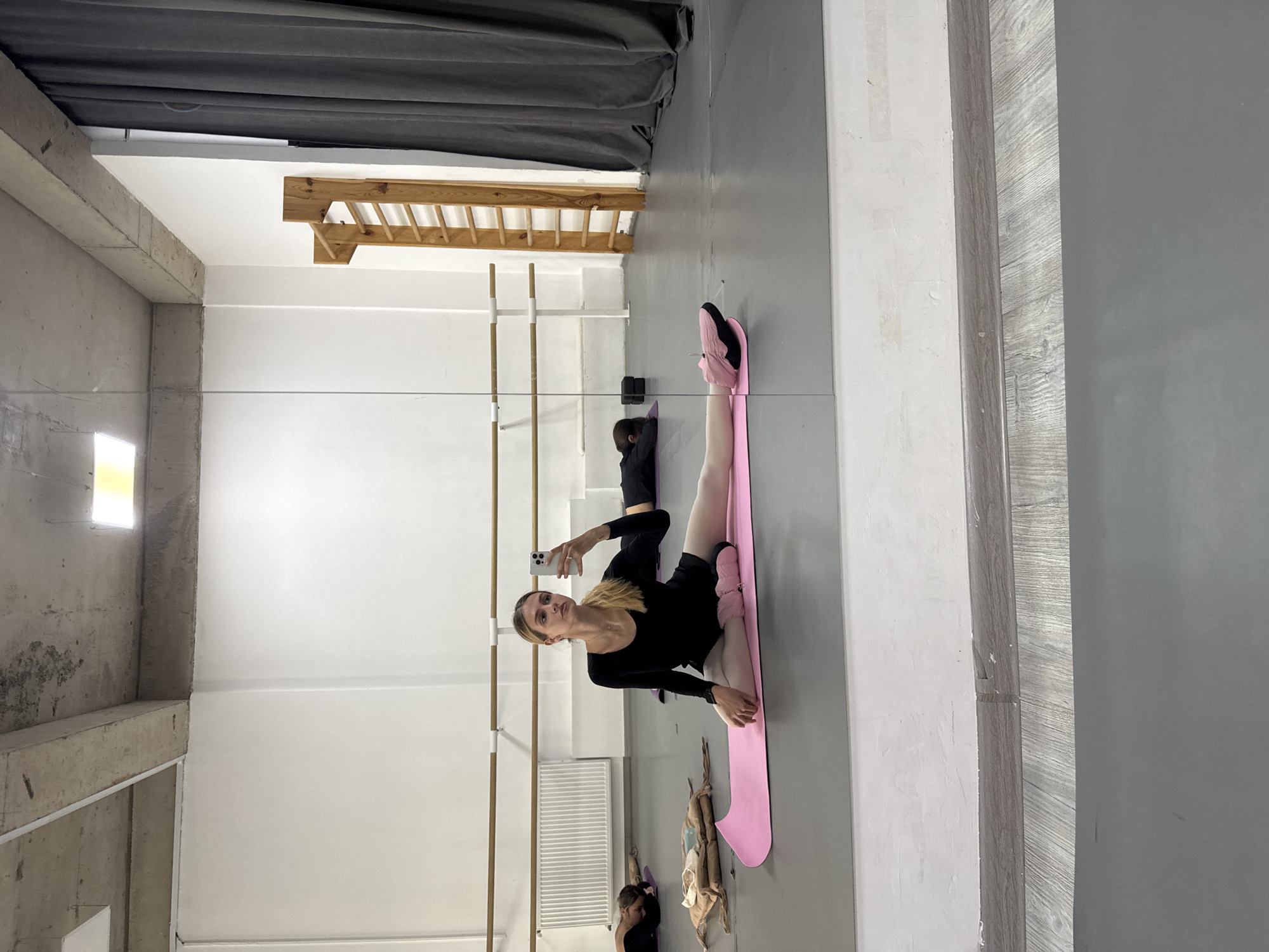 A photograph shows a woman doing a split on a pink mat in what appears to be a dance studio or gym. The woman is dressed in black leotard and leggings, with pink ballet slippers on her feet. She is facing a large mirror, holding a smartphone in her hand, seemingly taking a selfie. The reflection in the mirror shows a room with ballet barres along the wall, a curtain, and a window with a light source. The room seems spacious and utilitarian. The location in Tbilisi, Georgia, cannot be confirmed solely based on the image.