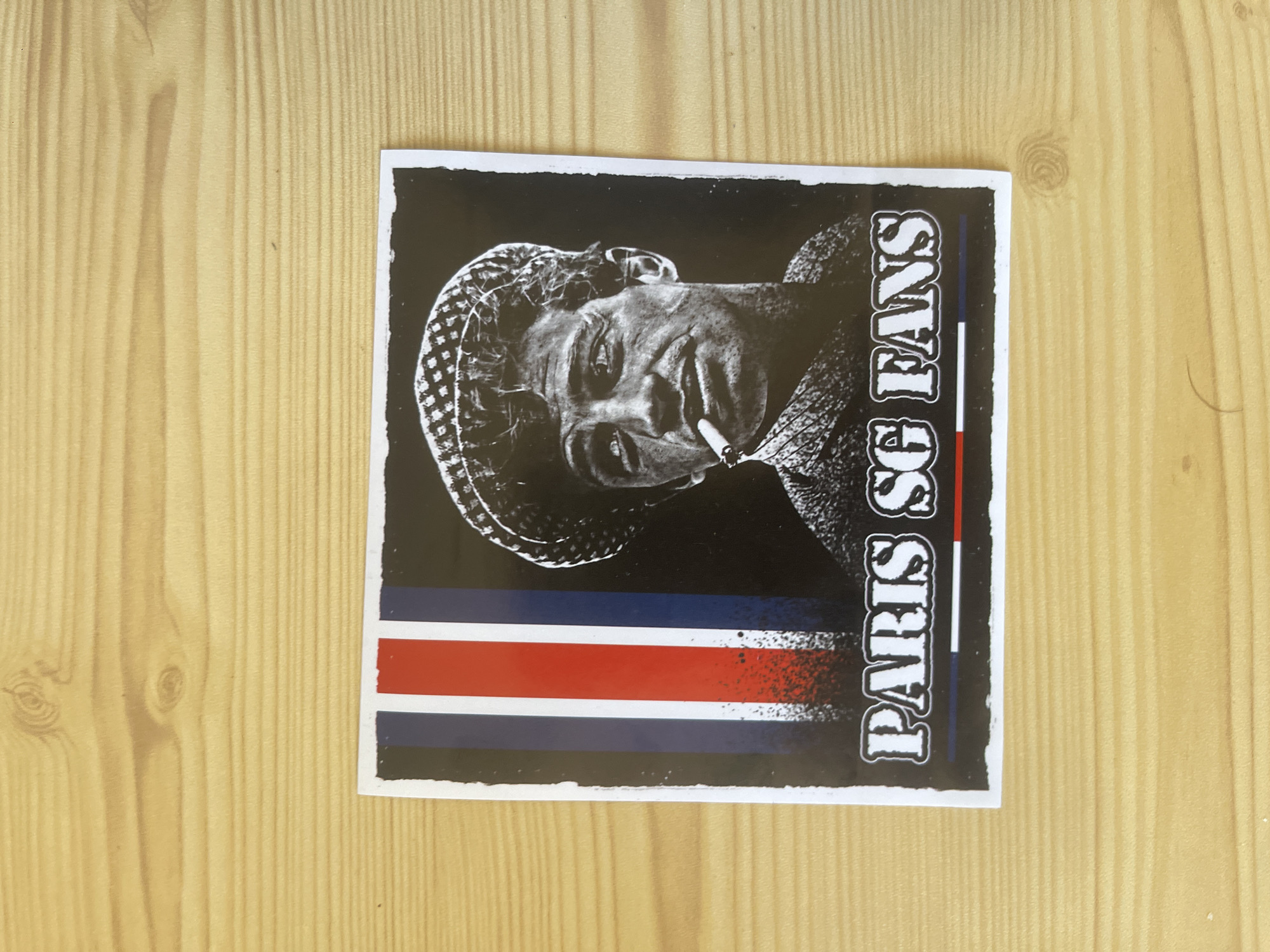 This is a sticker on a wooden surface. The sticker features a black and white image of a person's face with a cigarette in their mouth, and the text "PARIS SG FANS" written vertically on the right side. Below the face are three horizontal stripes: blue, red, and blue. The wooden surface appears to be a tabletop or floor, with visible wood grain. The orientation of the sticker suggests it is a portrait-oriented image. The context of "Paris SG FANS" implies a connection to Paris Saint-Germain Football Club.