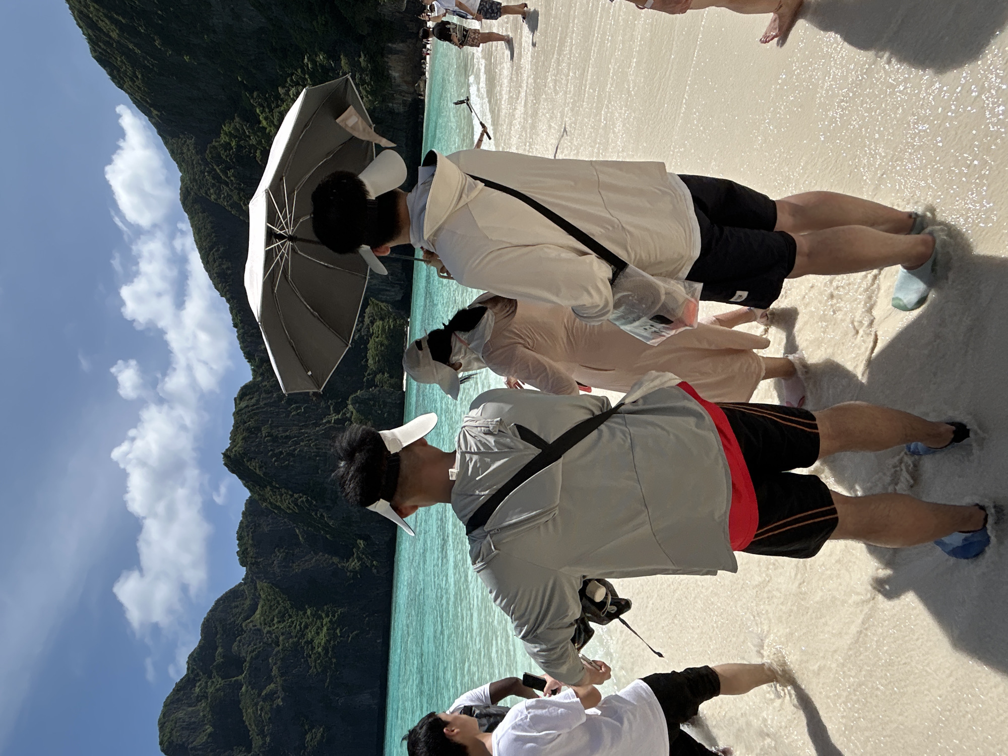 In Ao Nang, Thailand, a group of people stand in shallow, clear turquoise water on a sandy beach. Two men are prominently featured in the foreground, facing away from the camera. The man on the left wears a light gray hooded jacket over a red shirt and black shorts. The man on the right wears a cream-colored jacket and black shorts. Both men have black sling bags across their chests. Behind them, a woman in a light-colored dress and a cap is visible, holding an open grey umbrella above her head, seemingly to shield herself from the sun. Further in the background, other individuals are scattered along the beach and in the water. Towering, lush green cliffs rise dramatically behind the turquoise water, under a bright blue sky with scattered white clouds. The scene depicts a tropical beach setting with a group of tourists enjoying the water and scenery.