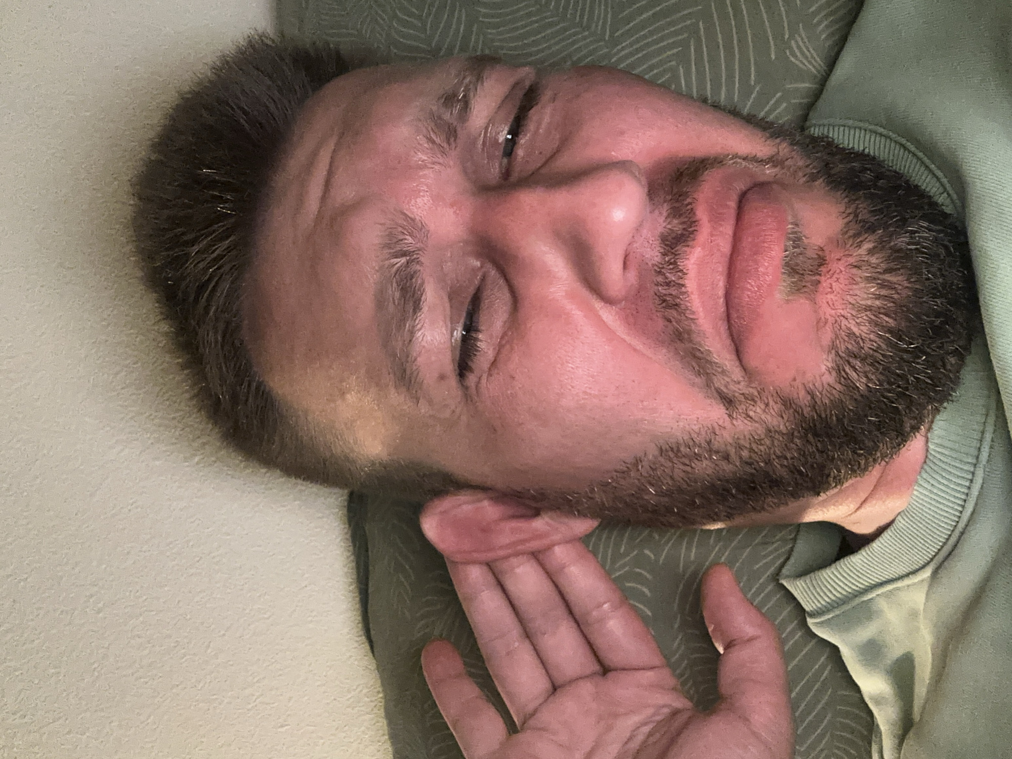 A close-up, low-angle, selfie-style shot shows a man's face from the nose up, with his right hand cupped behind his right ear. The man, who appears to be in his late 30s or early 40s, has a light complexion and stubble on his chin and upper lip. His hair is dark brown and short on the sides, longer and styled on top. He is wearing a light green crewneck sweatshirt. His eyes are partially closed, and his eyebrows are furrowed, suggesting a look of concentration or discomfort. The background consists of a textured wall and a patterned surface, possibly a pillow or cushion. The lighting is warm and somewhat dim, creating shadows on the man's face. The location is not specified in the image itself.