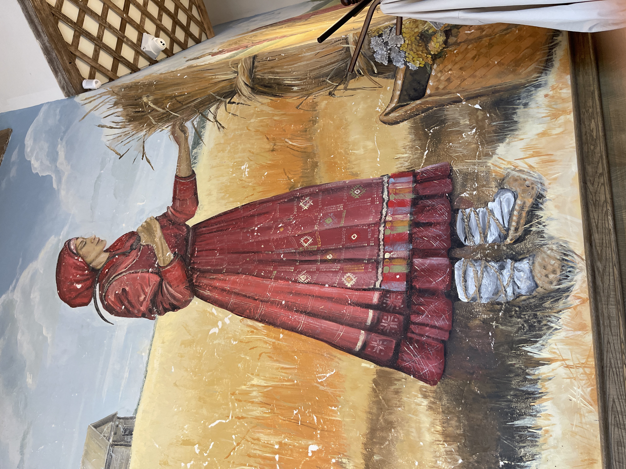The image shows a mural painting on a wall. The mural depicts a woman dressed in a traditional red garment, holding a bundle of wheat. The background consists of a field of wheat, a blue sky with white clouds, and a structure that could be a building or a barn. To the right of the woman is a basket filled with what appears to be yellow and silver flowers. The bottom part of the mural fades into darker tones. The mural seems to be framed. The location is unknown.