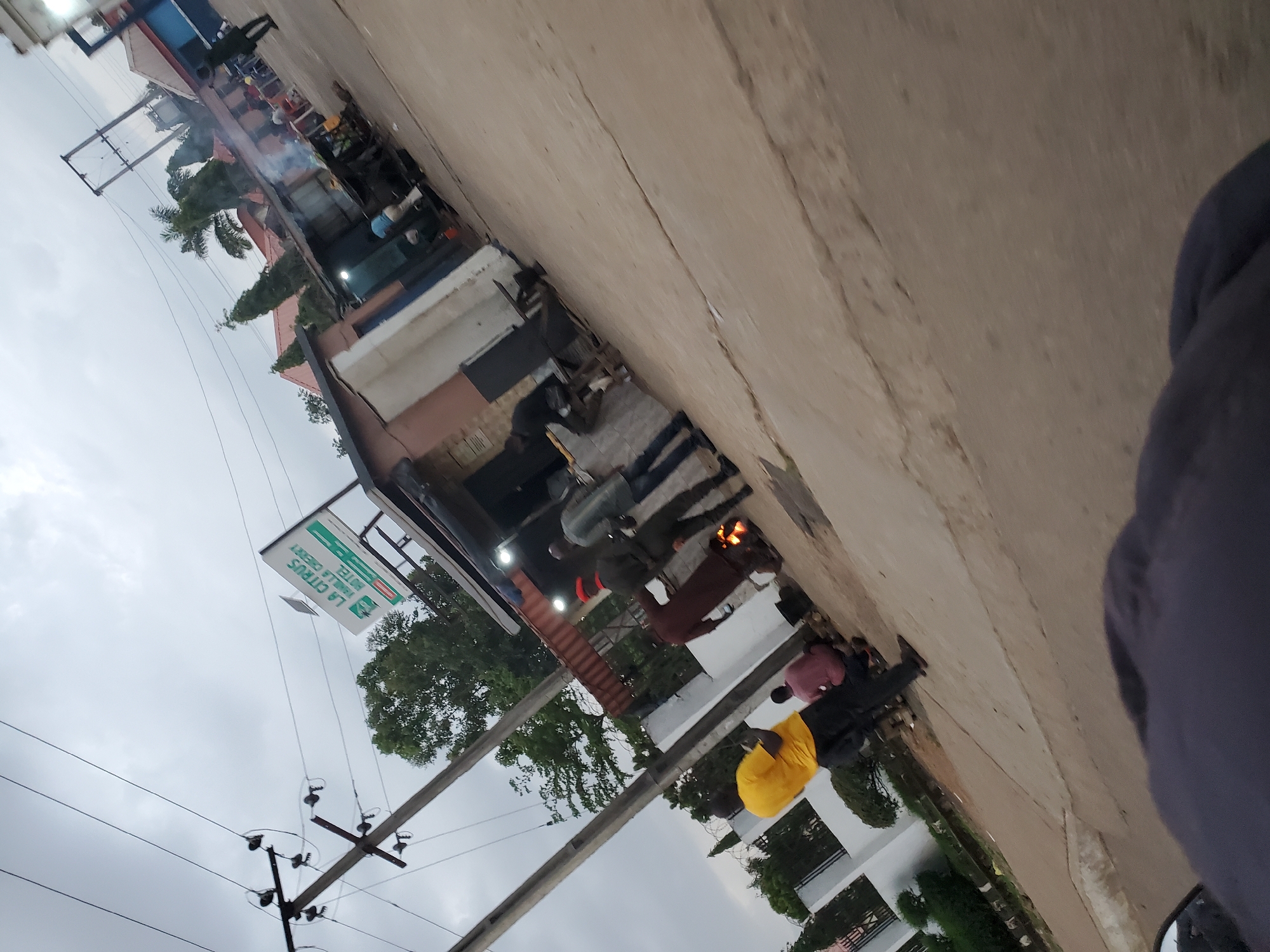 The image depicts an outdoor scene in Bukuru, Nigeria, likely along a street. In the foreground, a person wearing a yellow shirt and black pants stands facing away from the camera, with another person in a red shirt partially visible kneeling nearby. A fire burns in the middle ground, with several individuals gathered around it, some appearing to be cooking or preparing food. One man, wearing a dark uniform and a red cap, is seen near the fire. Behind the fire, a small building with a corrugated metal roof serves as a stall or shop. A sign with "LA CITRUS" and "HOTEL" in green lettering is visible above the shop. Power lines and utility poles are present in the background, along with trees and other buildings. The overall impression is a bustling street scene with food vendors and daily activities.