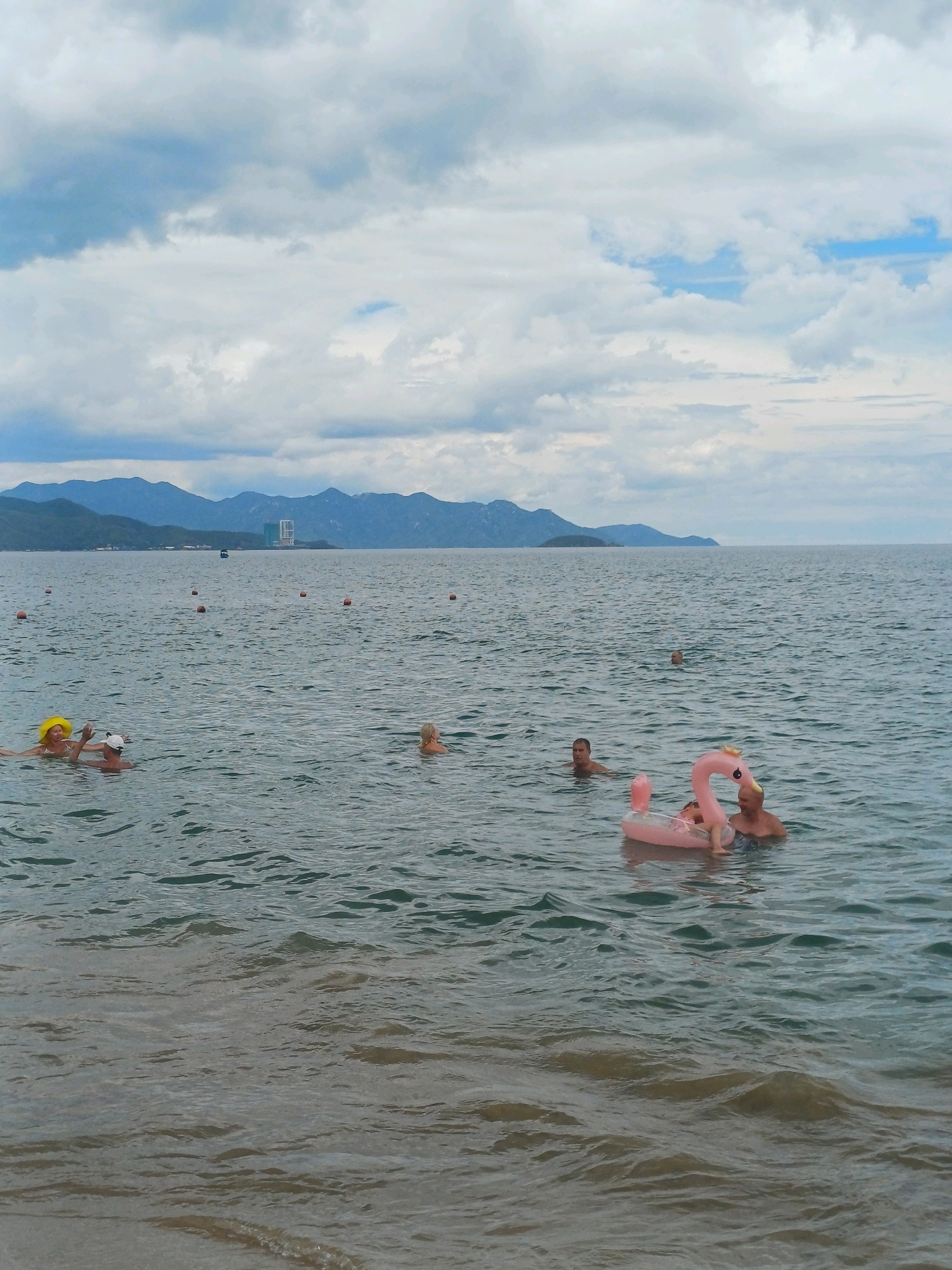 The image shows a body of water, likely the sea, with several people swimming. In the foreground, a pink inflatable flamingo float carries two individuals, an adult male and a child. Another adult male is visible nearby, partially submerged. To the left, two individuals, one wearing a yellow hat, are in the water. Further in the distance, another person is swimming. The water is choppy, and in the background, a coastline with mountains and some buildings can be seen under a cloudy sky. The scene is consistent with a public beach area in Nha Trang, Vietnam, possibly during daylight hours.