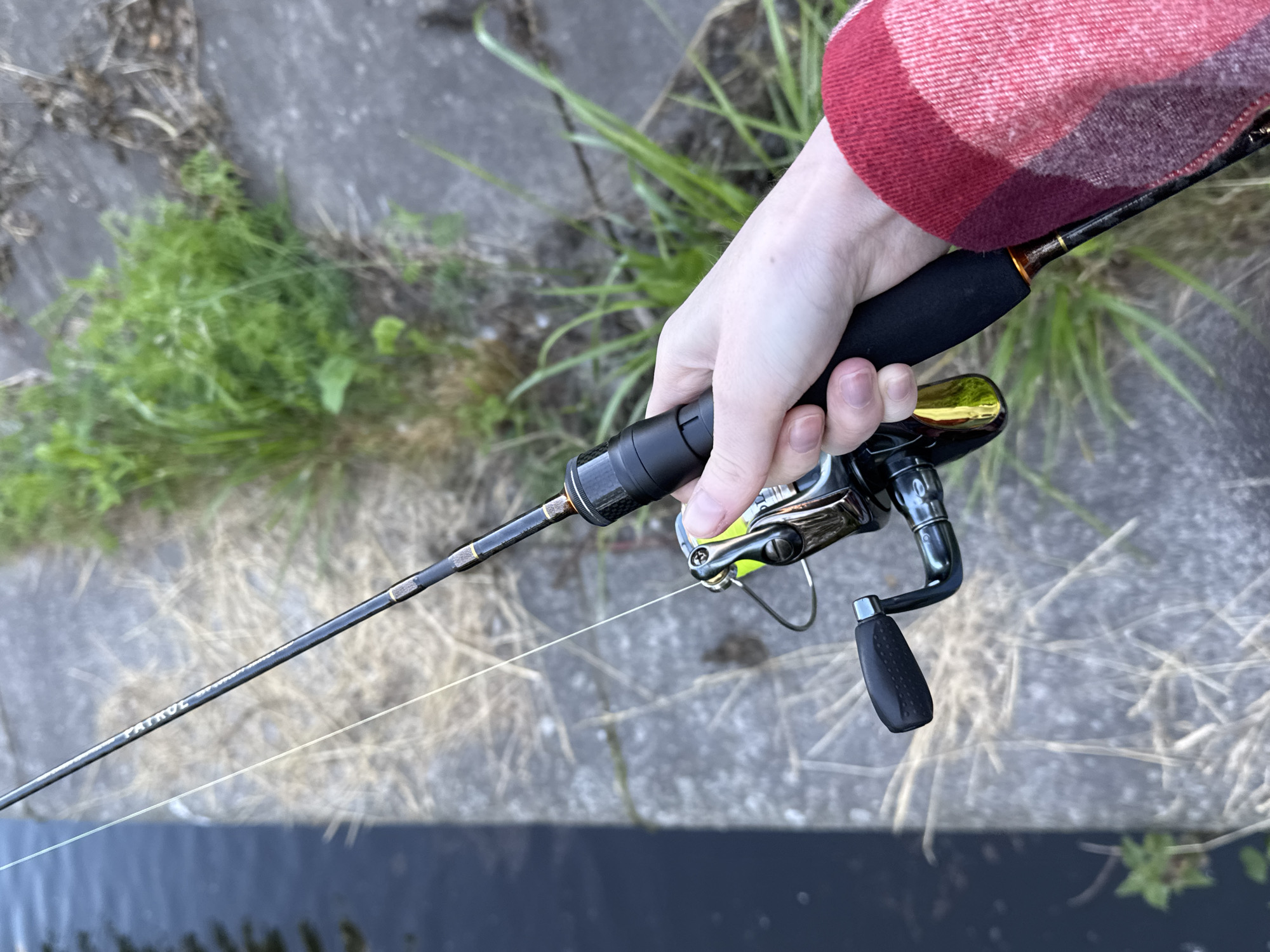 The image shows a close-up, high-angle shot of a person's hand holding a fishing rod and reel. The rod is black with gold accents and has the word "PATROL" visible on it. The reel is silver and black with a gold handle. The person is wearing a red and black plaid shirt sleeve. The background consists of a body of dark water on the left and a concrete embankment with grass and dry vegetation on the right. The scene appears to be outdoors, near a body of water, potentially a canal or river, in Saint Petersburg, Russia, given the context provided. The lighting suggests it is daytime. The focus is on the fishing gear and the hand holding it, with the environment slightly out of focus.