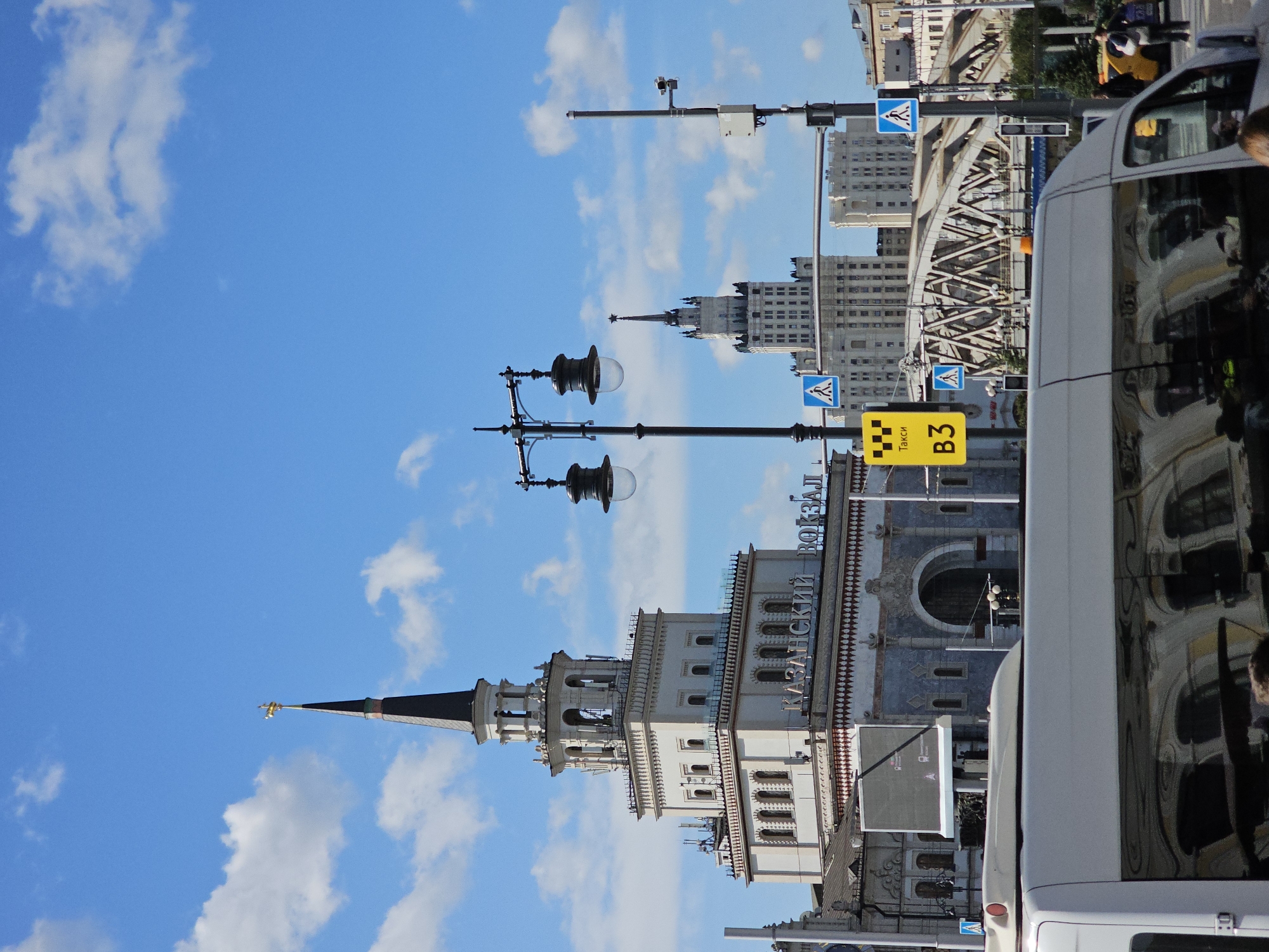 This is a vertical, medium shot captured in Moscow, Russia, likely during daytime, as indicated by the bright sunlight and blue sky with scattered clouds. The main subject is Kazan Railway Station, a prominent architectural landmark, with its distinctive white facade, ornate detailing, and a tall spire topped with a golden figure. The station's name, "КАЗАНСКИЙ ВОКЗАЛ" (Kazan Railway Station), is clearly visible on the building.
In the foreground, the top portion of a white van is visible, reflecting the surrounding buildings and sky. To the left of the van, a portion of a digital billboard is present. Two ornate lampposts with dark lanterns stand prominently in the midground, one closer to the foreground and another further back.
Beyond the station and the lampposts, another large, light-colored building with a pointed spire is visible, suggesting a Stalinist-era skyscraper typical of Moscow's architecture. A metal railway bridge with intricate truss work spans across the scene in the background. Traffic signals and pedestrian crossing signs are also present, indicating a busy urban environment.
A few people are visible in the far background near the yellow taxi and on the sidewalk, suggesting normal city activity. The overall scene depicts a bustling urban landscape centered around a historic railway station.