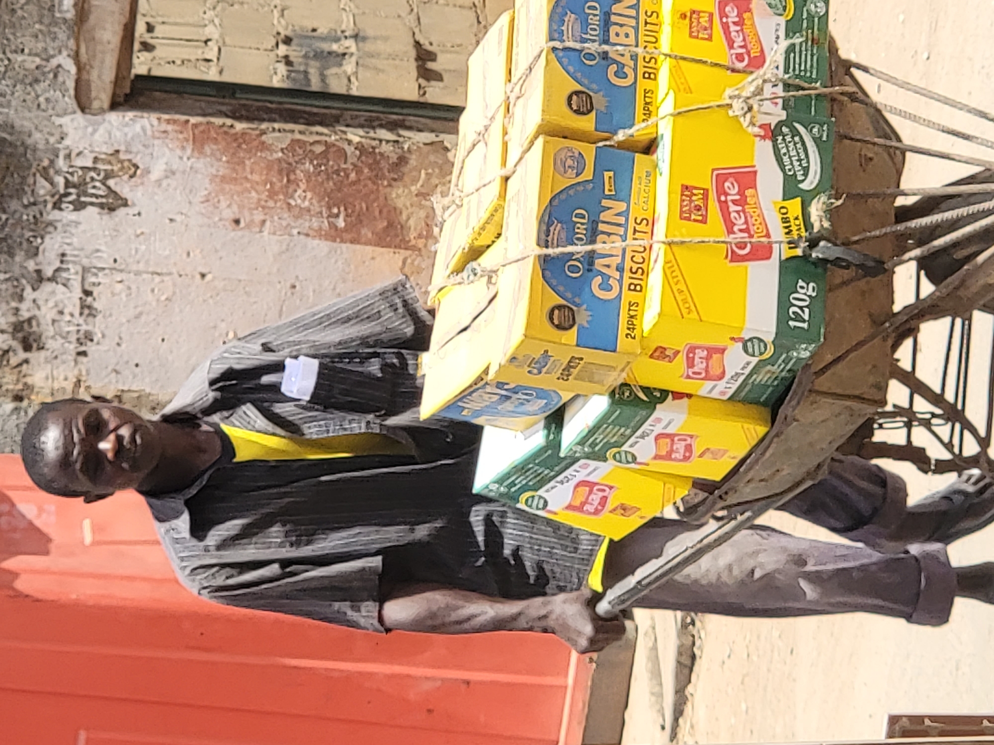 A man is captured in a full-body shot, standing next to a cart laden with packaged goods, likely on a street or market in Maiburiji, Nigeria. He is looking directly at the camera with a neutral expression. The man wears a striped dark grey shirt with a yellow element visible at the collar, and dark grey trousers. His left arm is visible, holding onto the cart's frame, while his right arm is mostly obscured by the boxes.
The cart, a simple two-wheeled pushcart, is heavily loaded with multiple yellow and green boxes, securely tied with rope. The background features a distressed reddish-brown brick wall with some white plaster visible, suggesting an urban or semi-urban outdoor setting. Bright daylight illuminates the scene, casting soft shadows, indicating it is daytime.
Visible text on the boxes includes product names and details: "OXFORD CABIN 24PKTS BISCUITS CALCIUM" appears on several yellow boxes. Other packages, green and yellow, display "Cherie noodles", "TASTY TOM", "LUMBO PACK", "CHICKEN PEPPERSOUP FLAVOUR", and "120g". These products appear to be biscuits and instant noodles, indicating the man is likely a vendor or transporter of these goods.