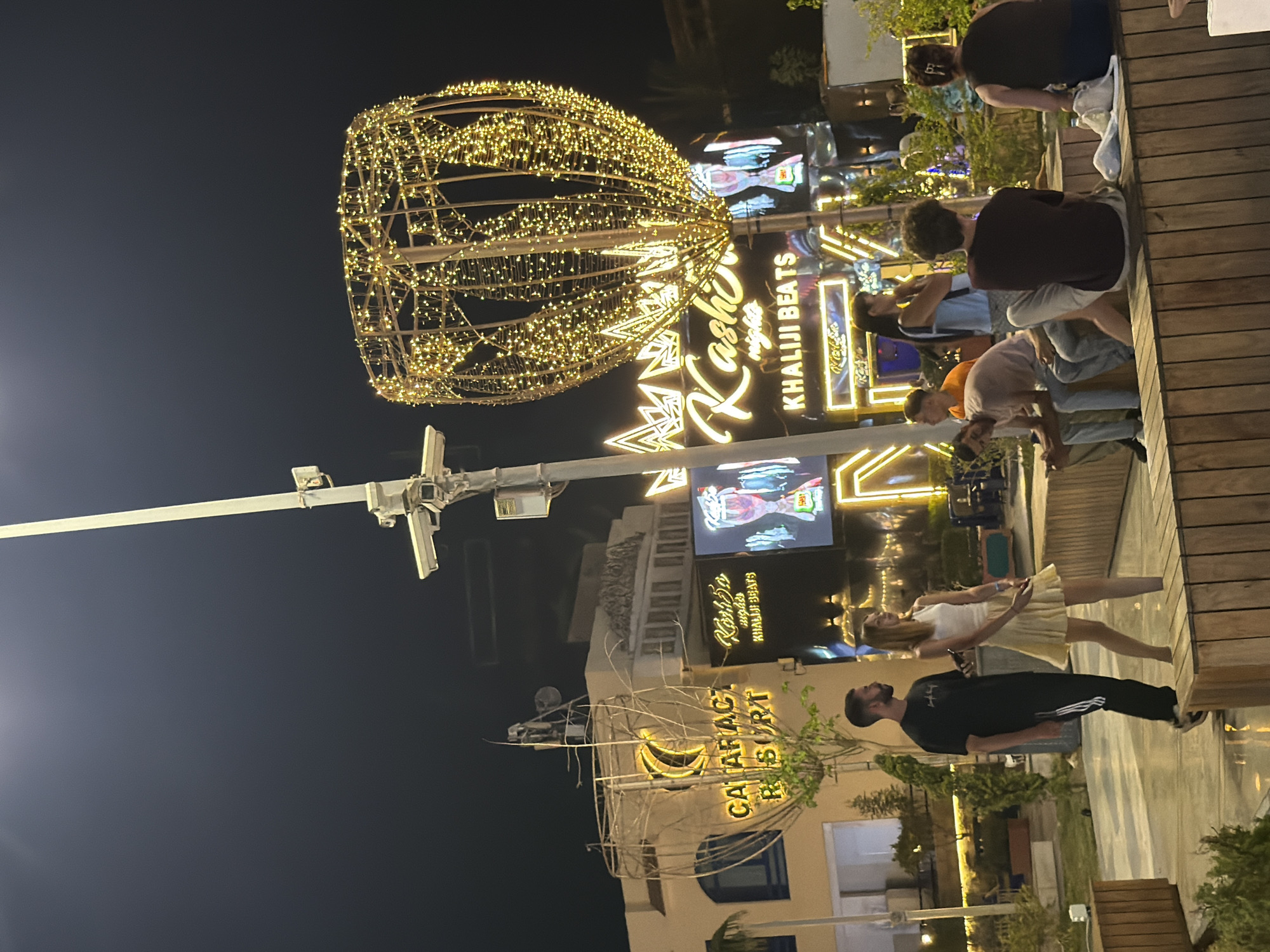 In Sharm El Sheikh, Egypt, an outdoor evening scene unfolds. A group of young adults are gathered in a public space, possibly a promenade or seating area, illuminated by decorative lighting. In the background, a prominent building displays signage for "Cataract Resort" and "Kashka nights KHALIJI BEATS." A large, elaborate light fixture, shaped like an oversized bulb or flower bud and adorned with numerous small, warm-toned lights, dominates the upper right portion of the frame, casting a festive glow. Another, more abstract wireframe structure, also decorated with branches and lights, stands to the left. Several people are seated on wooden benches, while others are standing, interacting or observing their surroundings. A man and a woman are standing in the mid-ground, facing away from the camera. The ground appears to be paved, with some areas reflecting the ambient light, suggesting it may be wet. Lush green vegetation and trees are visible around the seating area and in the background. The overall atmosphere is one of leisure and entertainment, typical of a tourist destination at night.