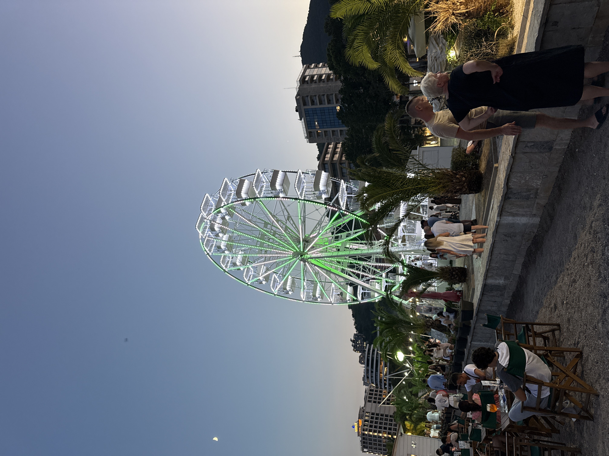 An outdoor evening scene is depicted in Budva, Montenegro, near the coast. In the foreground, a gravel area with several tables and chairs is set up, suggesting an outdoor dining or seating area. Some people are seated at the tables, some appear to be waiting, and others are walking by. A large Ferris wheel dominates the mid-ground, illuminated with bright green and white lights. Palm trees are scattered throughout the scene, contributing to a tropical or coastal atmosphere. In the background, several multi-story buildings and a hill with some structures are visible against a twilight sky. A crescent moon is faintly visible in the upper left quadrant of the sky. The overall impression is of a lively evening in a resort town.