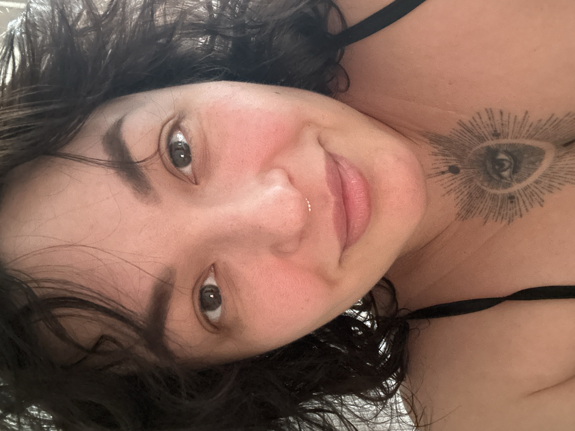 The image displays a close-up, frontal portrait of an adult female from the chest upward. She has dark, curly hair, brown eyes, and fair to medium skin tone with visible blush on the cheeks. A small, metallic septum piercing is present above her upper lip. On her upper chest, centrally located, is a black ink tattoo depicting an eye within a radiating sunburst pattern, surrounded by several small dots. A black clothing strap is visible over her right shoulder. She maintains direct eye contact with the viewer and exhibits a slight smile. The background is out of focus, appearing as an indistinct light-colored surface, indicative of an indoor setting. No specific location within Chaikovsky, Russia, or any other geographical detail is discernable from the visual evidence in the image.