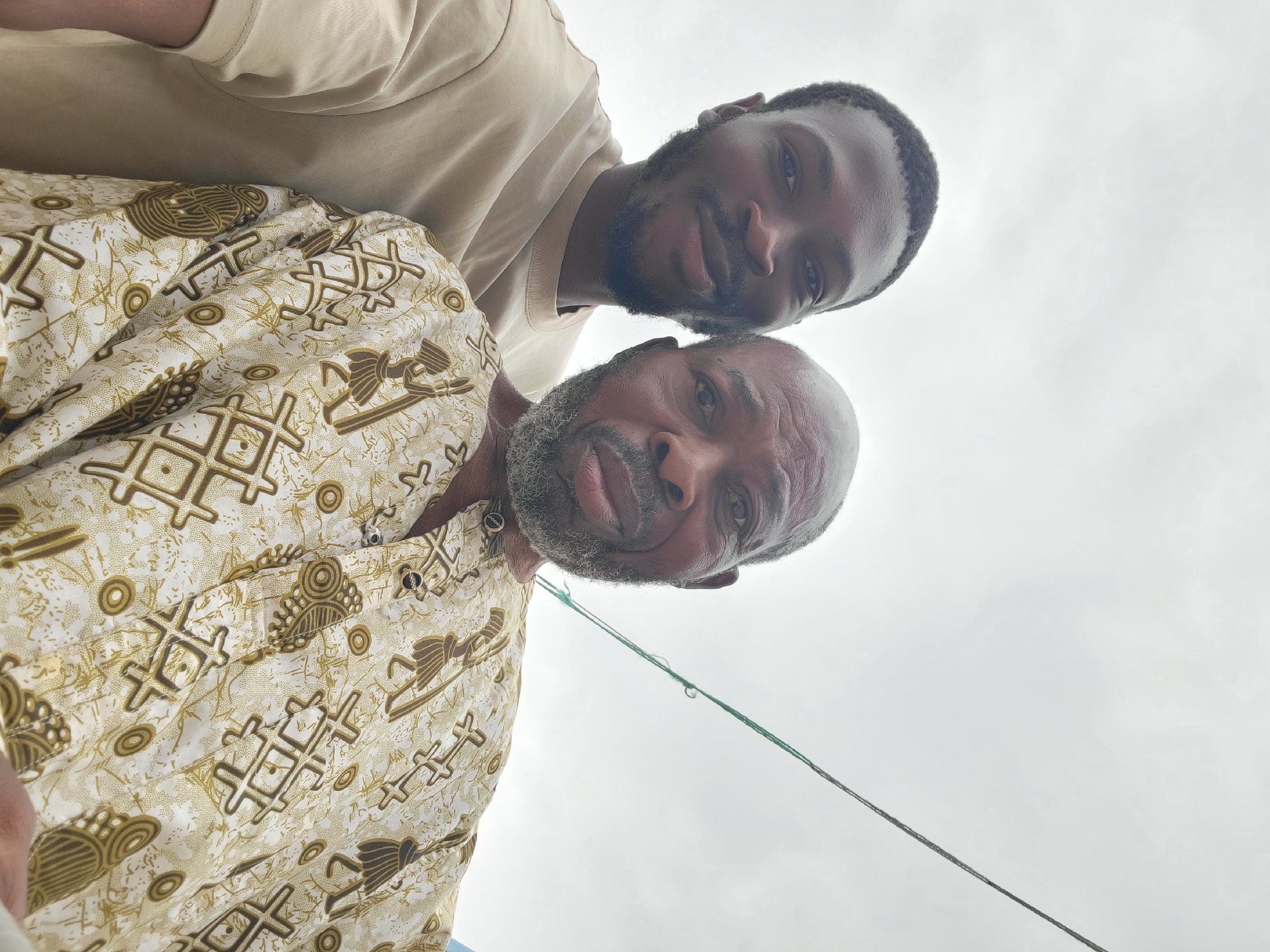 Two men, one younger with dark skin and short black hair and beard, and an older man with dark skin, a bald head, and a graying beard, are pictured from the chest up in a selfie. The younger man is on the left, wearing a tan crewneck shirt. The older man is on the right, wearing a patterned gold and white shirt with brown symbols resembling stick figures and geometric shapes. A thin green and gray rope extends diagonally across the upper right quadrant of the frame. The background is a cloudy, overcast sky.