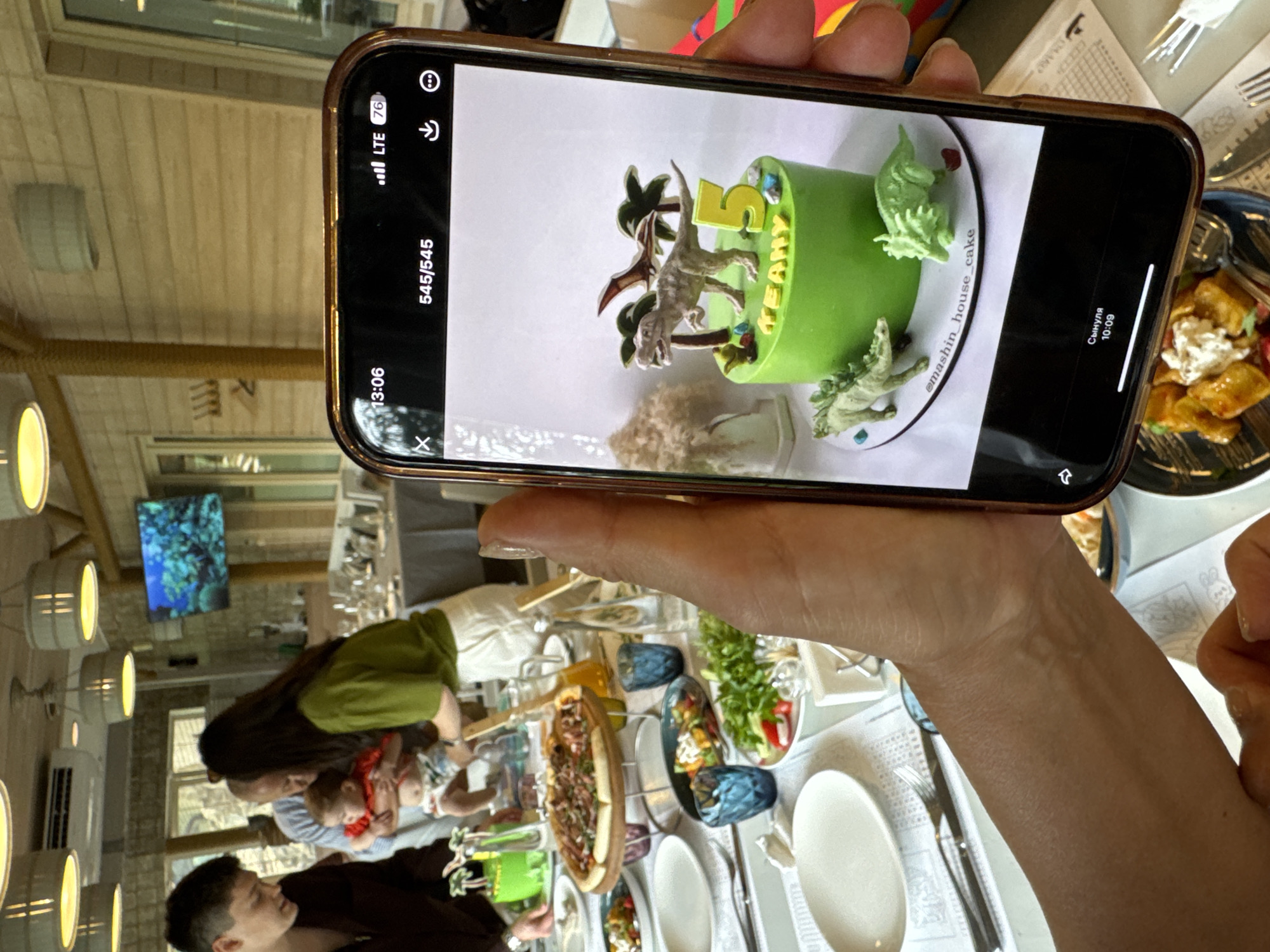 The image depicts a celebratory scene at a restaurant, likely in Sovkhoz imeni Lenina, Russia, as evidenced by the text on the phone. A phone is held in the foreground, displaying a photo of a dinosaur-themed cake. The cake is bright green with toy dinosaurs and palm trees as decorations, and the name "KEAHNY" is written on it in yellow lettering, along with the number "5". The phone shows the time as 13:06 and the image number as 545/545.
In the background, blurred figures are visible around a table set for a meal. There is a pizza on a wooden board, salads, and drinks on the table. A baby is being held by an adult. The restaurant appears to have a rustic or natural aesthetic, with wooden walls and overhead lighting. A television screen displaying an underwater scene is mounted on the wall in the distance. The overall atmosphere suggests a birthday celebration.