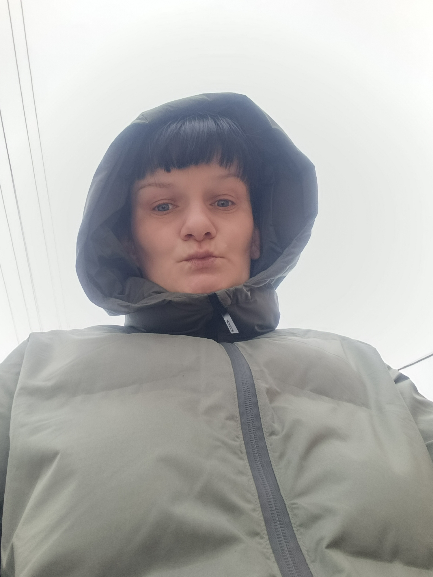 A low-angle close-up shot depicts a woman in a green hooded jacket against an overcast sky. The woman is centered in the frame, with the hood framing her face and the jacket filling the lower portion of the image. Overhead electrical wires are visible in the upper left of the frame. Based on prior knowledge, the location is specified as Фоки, Russia.
