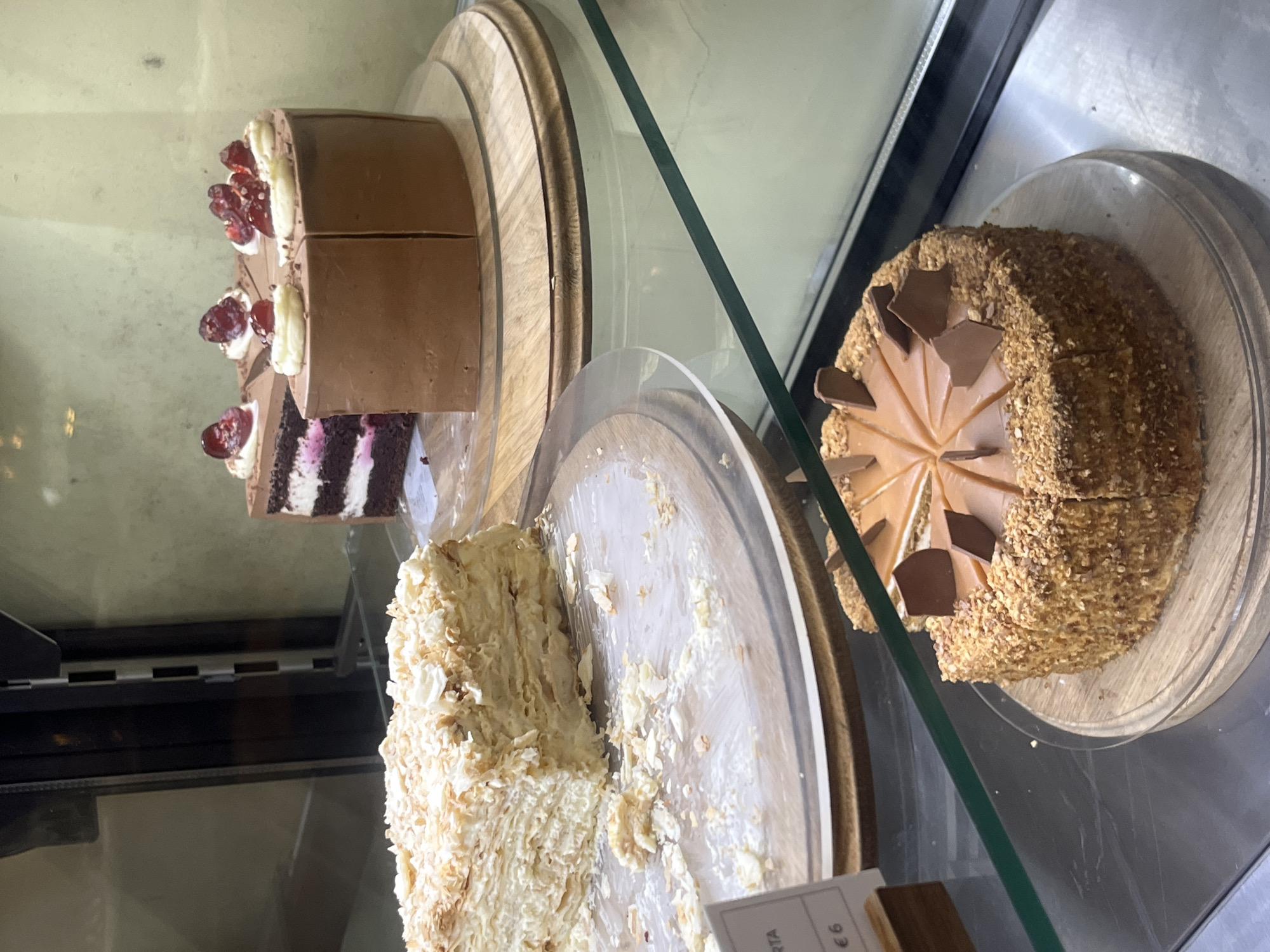 The image displays the interior of a commercial refrigerated display case showcasing multiple cakes, located in Bečići, Montenegro.
On the uppermost visible shelf, two round chocolate cakes are presented on a light-colored wooden circular stand. The cake on the left has a significant portion removed, revealing internal layers of dark chocolate cake, white cream, and a reddish fruit filling. Its top is decorated with white cream dollops and whole red berries. The cake on the right, largely intact, is similarly decorated with white cream dollops and red berries.
The middle shelf features a large, rectangular section of a multi-layered cream cake, characterized by numerous thin, flaky pastry layers and white cream filling. A substantial portion of this cake has been removed, leaving a scattering of light-colored crumbs on its clear circular display plate. A white price label, partially visible on the left, displays "TA" and the price "€6."
On the lowest visible shelf, a round cake with a granulated or textured light brown exterior, likely coated in nuts or crumbs, is displayed on a light-colored wooden circular stand. Its top is covered with a smooth, light brown glaze and adorned with several dark brown, irregularly shaped chocolate shards. This cake has been cut into radial wedges, with multiple individual slices already removed.
The display case itself is constructed with reflective glass shelves, some exhibiting a green tint on their edges, and features dark vertical structural elements in the background. No individuals are visible within the scene.