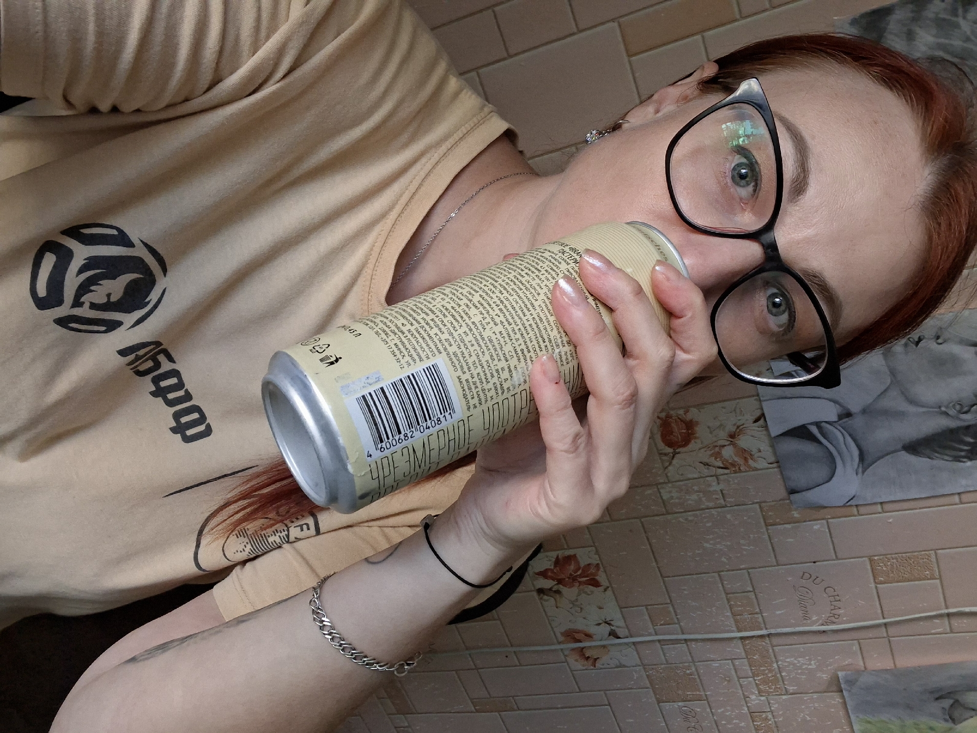 A woman with red hair and glasses is taking a drink from a can. She is wearing a light brown t-shirt with a black logo. She has a silver necklace and a silver bracelet. She is in front of a wall with patterned wallpaper. There are two black and white drawings on the wall. The drawings appear to be of people. The wallpaper has a tiled pattern and some floral designs. The scene is illuminated by artificial light.
