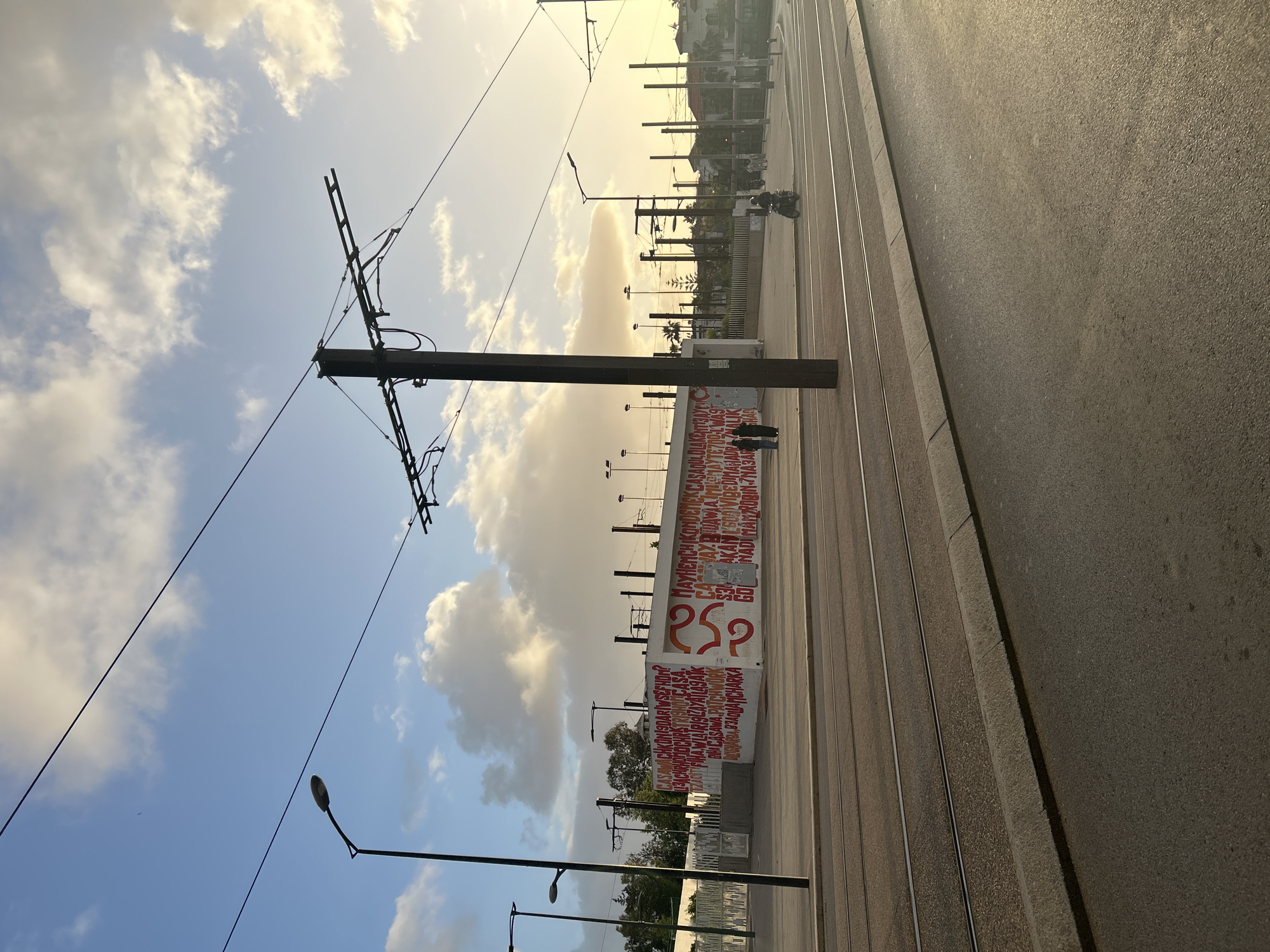 The image captures an outdoor scene in Casablanca, Morocco, near a light rail line. The photograph is taken near the street level, with the road taking up the right side of the frame. The light rail tracks run parallel to the road and are separated by a narrow concrete barrier.
A concrete wall runs parallel to the tracks, adorned with large, red lettering over a white background; parts of the text include "CASA", "CASAWA", and a large "RSP". Street lights and power lines stretch overhead, adding to the urban landscape.
The sky is visible, displaying a mix of blue and clouds. In the distance, trees and buildings are visible against the bright light. A scooter is also seen riding along the train tracks in the distance. Two people are standing near the wall.