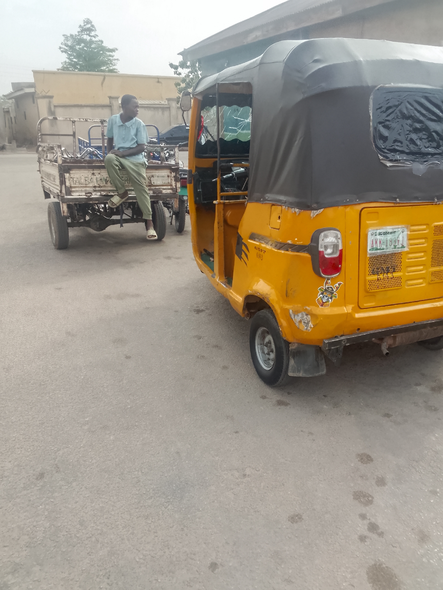 A young man sits on the back of a small, flatbed, two-wheeled trailer attached to a small, three-wheeled vehicle in Pantame, Nigeria. The trailer is white and has lettering on the back. The vehicle is yellow with a black vinyl top and a license plate that reads "COMBE AKK 1831". The street is paved and appears to be residential. Buildings are visible in the background. The time of day seems to be daylight, but overcast.