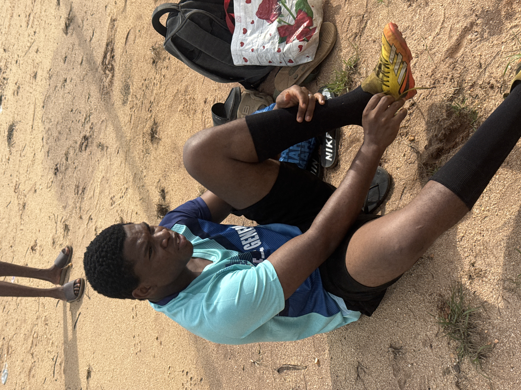 In Ihiagwa, Nigeria, a young Black male sits on sandy ground, tying the laces of a yellow and orange soccer cleat. He wears a light blue and dark blue athletic shirt, black shorts, and black socks. Next to him are a black backpack and a white tote bag decorated with roses and hearts. Another person's legs and feet, clad in sandals, are visible in the background. The environment is an outdoor sandy area with sparse vegetation.