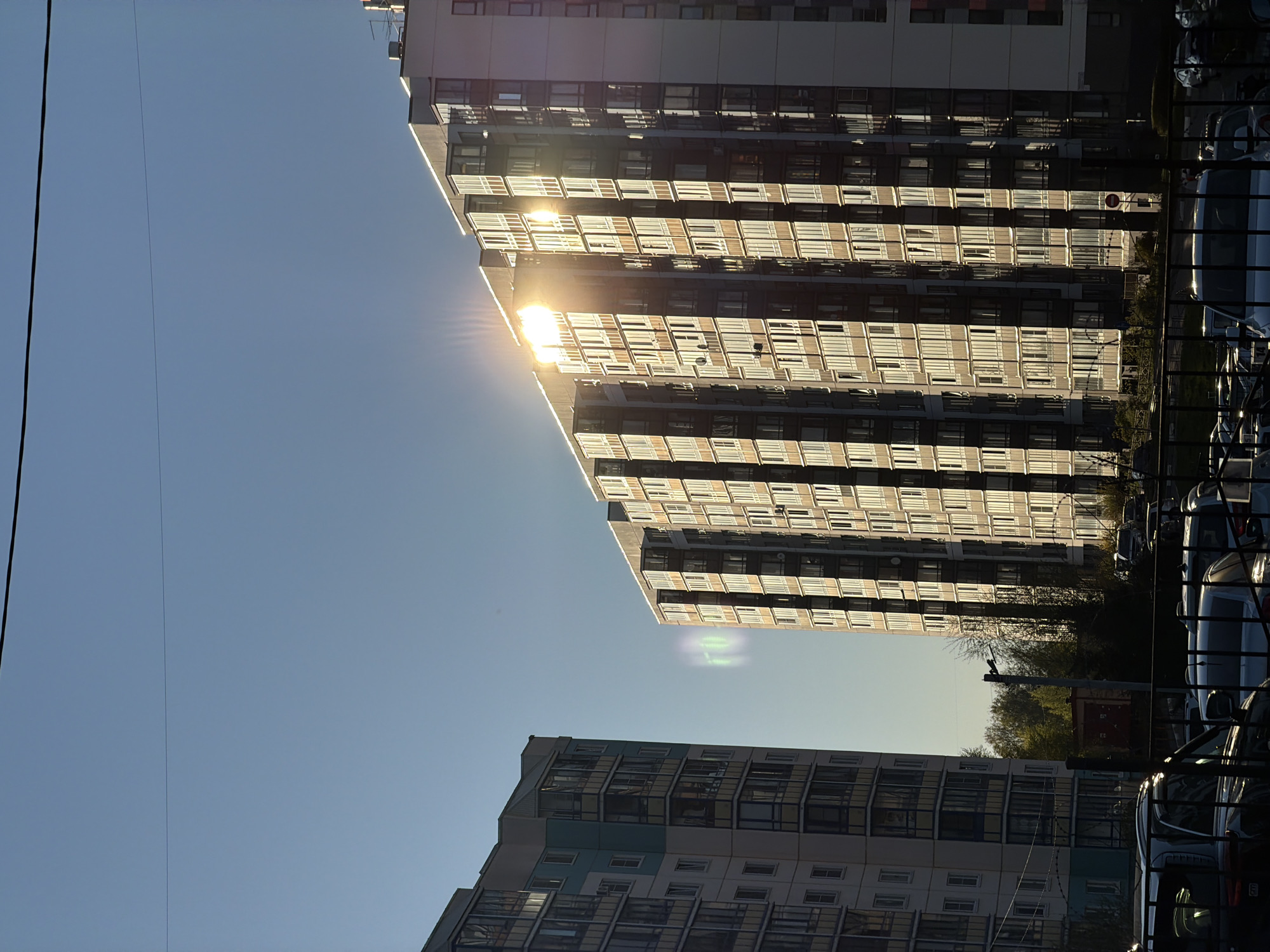 The image shows two residential buildings in an urban setting, likely in an Eastern European or Russian city based on the architectural style. The sky is clear and blue, and the sunlight reflects intensely off the windows of the taller building. Cars are parked along the street in front of the buildings. Power lines are visible against the sky. The photo is taken at a low angle, looking upwards. The buildings are modern, high-rise structures with multiple floors and many windows. The scene suggests a typical residential area in a city with dense housing.