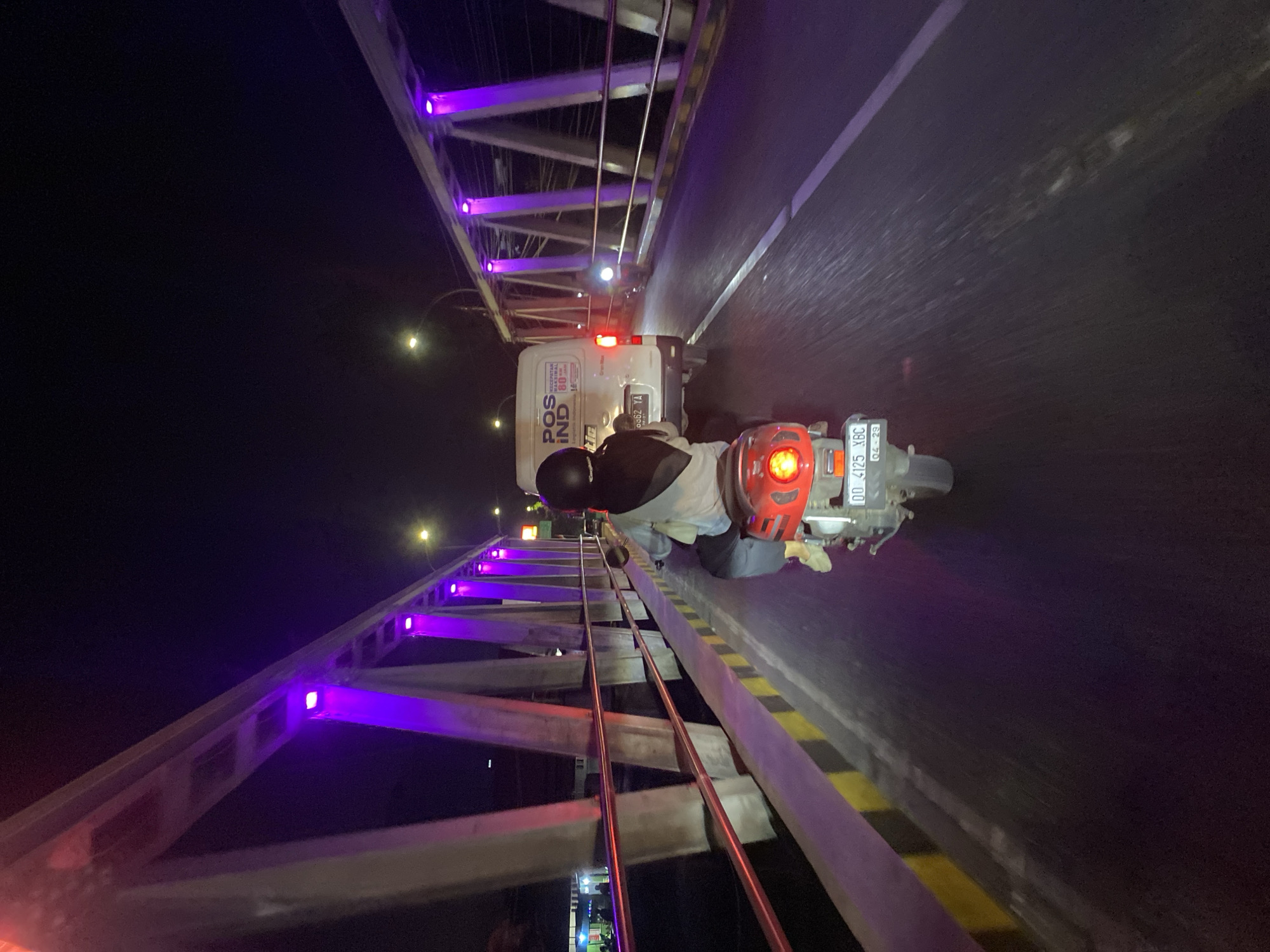 The image depicts a nighttime scene on a bridge in Bantaeng, Indonesia. A person wearing a black helmet and headscarf rides a red scooter on the bridge. The scooter has a license plate with the text "DD 4125 XBC 04-29". Ahead of the scooter, a white van with "POS IND" branding is visible. The bridge is illuminated by purple lights and streetlights. The road is wet, suggesting recent rain. The perspective is from behind the scooter, looking forward.