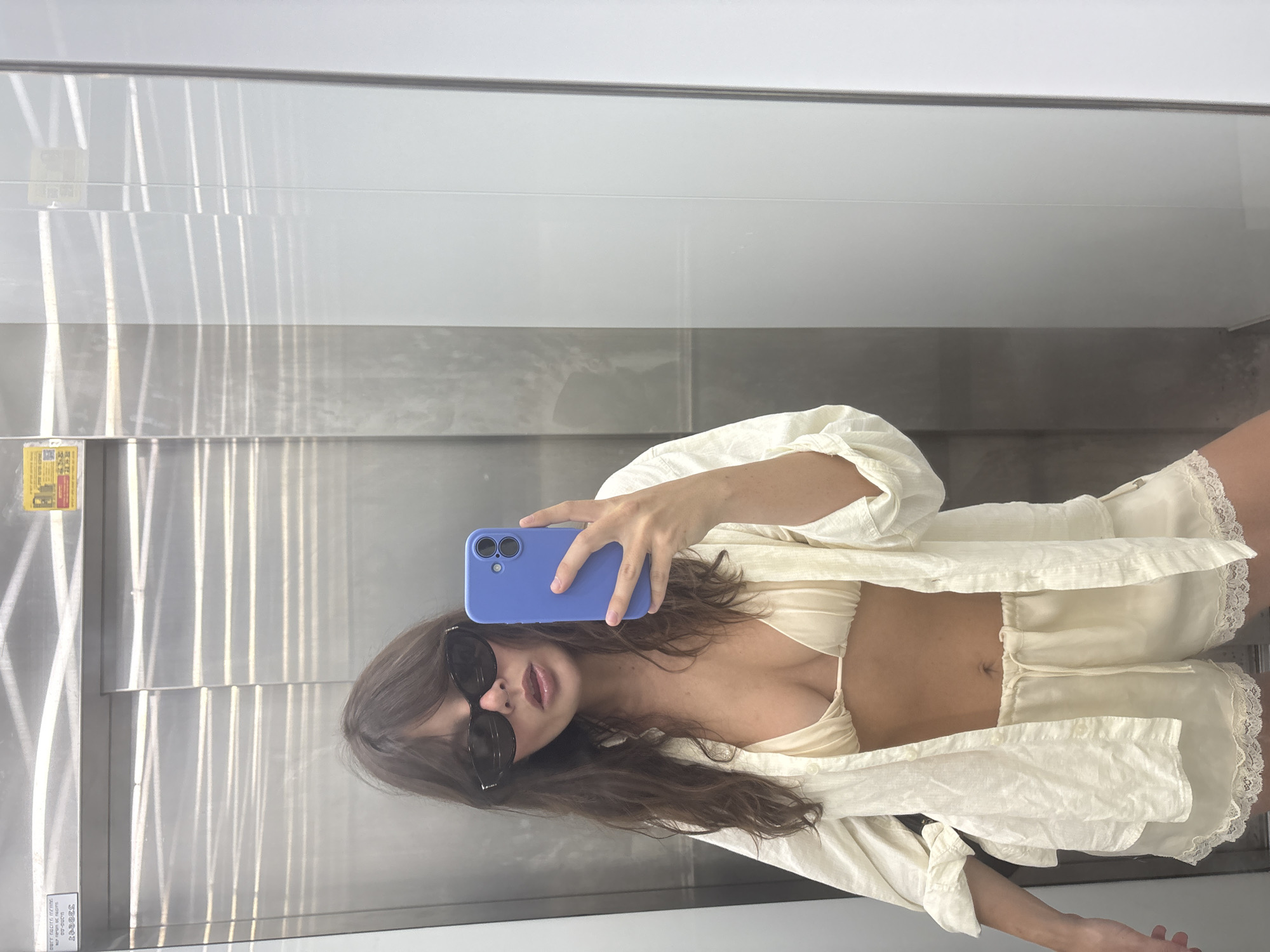 A young woman with brown hair and sunglasses is taking a mirror selfie in an elevator. She is wearing a cream-colored bikini top, an unbuttoned oversized cream-colored linen shirt, and matching cream-colored silk shorts with lace trim. She is holding a blue-cased smartphone in her right hand. The elevator has brushed stainless steel walls and bright fluorescent lighting. A yellow safety sticker is visible on the upper left wall of the elevator. The elevator appears to be located in Tel Aviv, Israel.