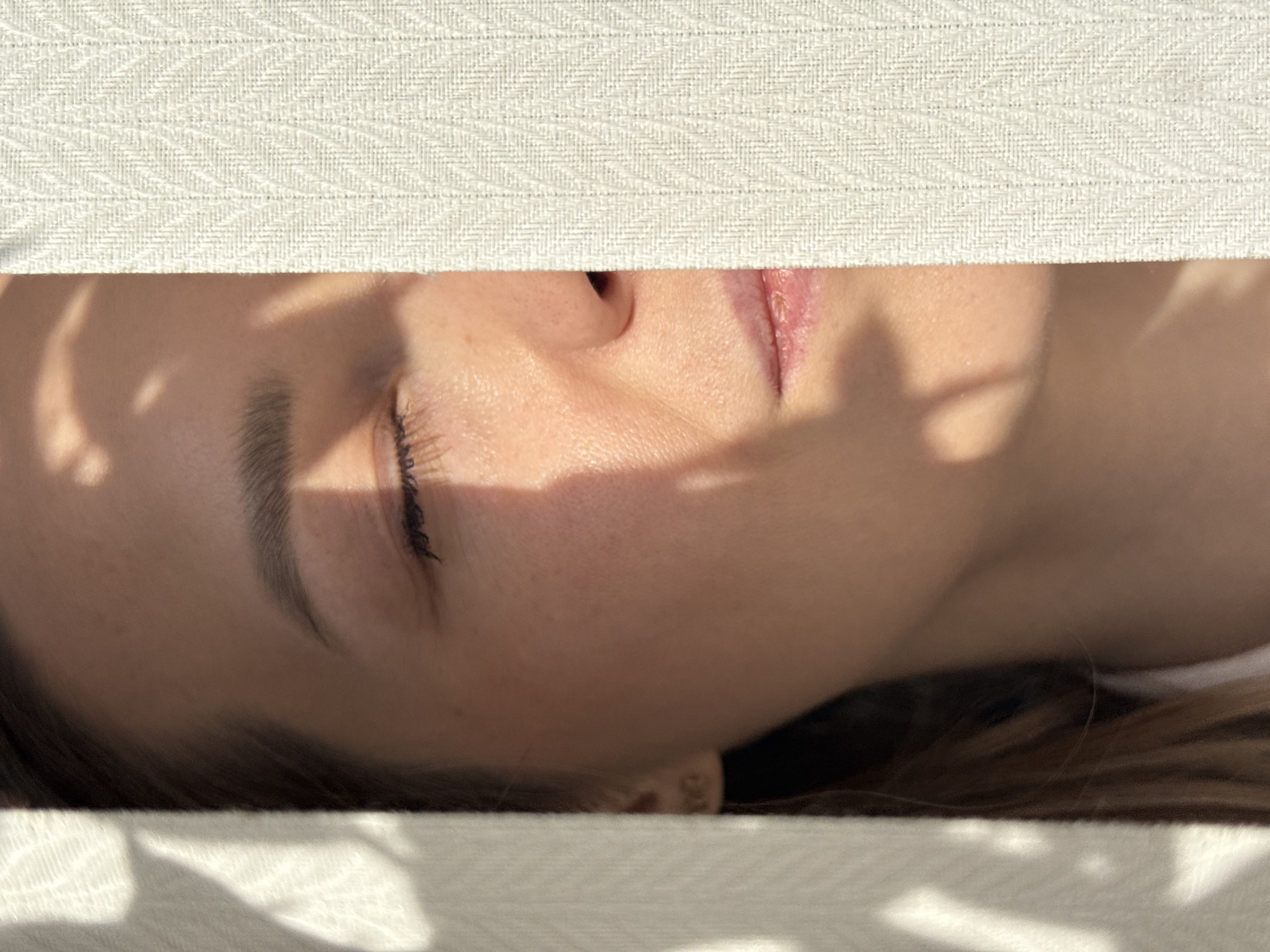 The image shows a person's face partially visible through a narrow opening between two light-colored, textured surfaces, possibly blinds or fabric. The person is lying down, with their eyes closed and their face partially obscured by shadows, possibly from foliage outside the opening. Details visible include an eyebrow, eyelashes, nose, and a portion of the mouth. The person's skin tone is light. The background and surrounding environment are not clearly visible. There is no information in the image or provided context that indicates the location is Chaikovsky, Russia.