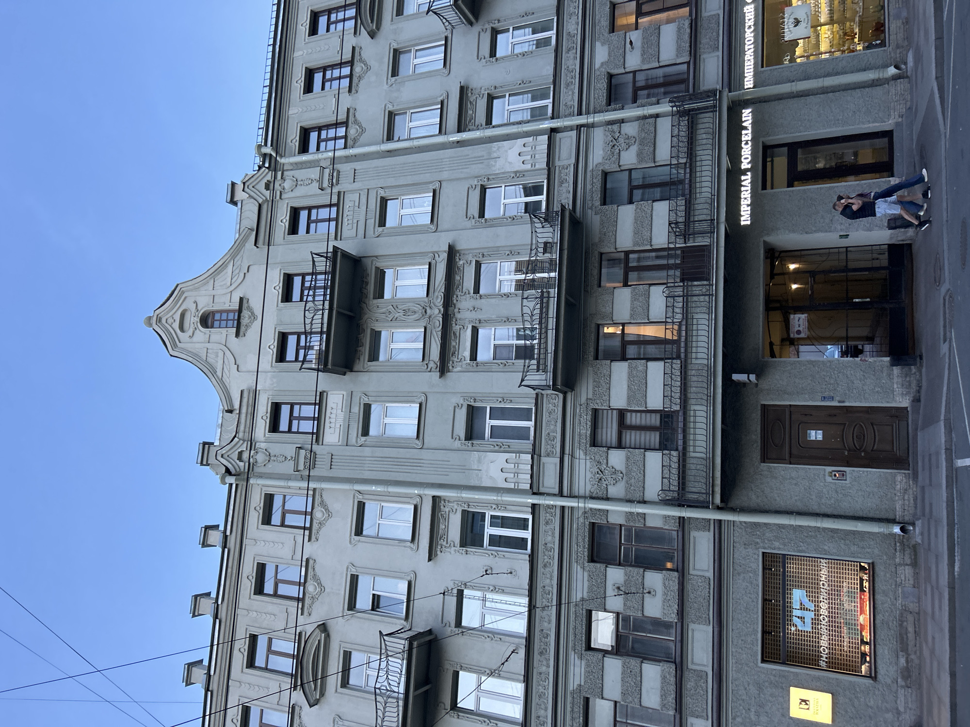 This is a medium shot, eye-level, exterior, vertical, full-color photograph of an old, ornate building with multiple stories and windows. The building is grey with intricate decorative detailing around the windows and balconies. There are several balconies with wrought-iron railings. The ground floor has storefronts. One storefront is for "IMPERIAL PORCELAIN" and another displays goods behind a window. Two people are walking on the sidewalk in front of the storefronts, one in a black shirt and blue shorts, the other in a purple shirt and blue jeans. The sky is clear and blue. There are power lines crossing the sky above the building. The scene is likely in a city given the building and sidewalk.