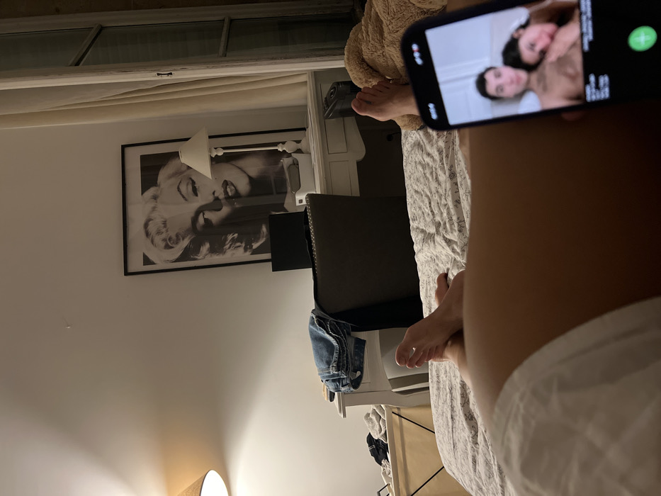 A person lies on a bed, holding a smartphone displaying an incoming video call with two men embracing. An oversized framed portrait of Marilyn Monroe hangs on the wall behind a desk with a lamp. A pair of jeans are draped over the desk. The room appears to be dimly lit. A window with partially drawn curtains is visible at the top of the frame. The bed has white and gray patterned linens.