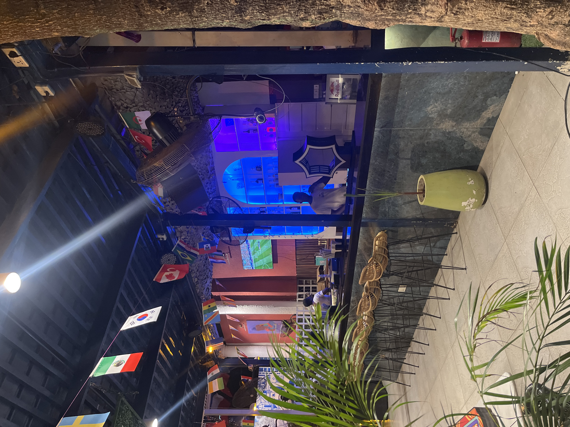 The image depicts an outdoor seating area of a bar or restaurant, likely in Accra, Ghana, as suggested by the presence of multiple national flags, including those of Ghana and Nigeria, among others. The scene appears to be set in the evening or at night, with artificial lighting illuminating the space. Blue and purple lights are prominent, creating a vibrant atmosphere around the bar area.
Several objects are visible, including a large fan, a television screen displaying a sports match, numerous national flags strung across the ceiling, and a red fire extinguisher. There are also several tables and chairs, with some wicker chairs lined up along a wall. A green vase with a plant stands on a tiled floor. The background shows a bar interior with shelves stocked with bottles.
There is one person clearly visible, a man with dark skin wearing a white t-shirt, sitting at the bar. Other individuals are dimly visible in the background, suggesting the venue is moderately busy. The overall mood seems relaxed and social, typical of a bar environment, with the sports broadcast on the TV likely attracting patrons. The decor with the flags suggests a focus on international events, perhaps a sports tournament.