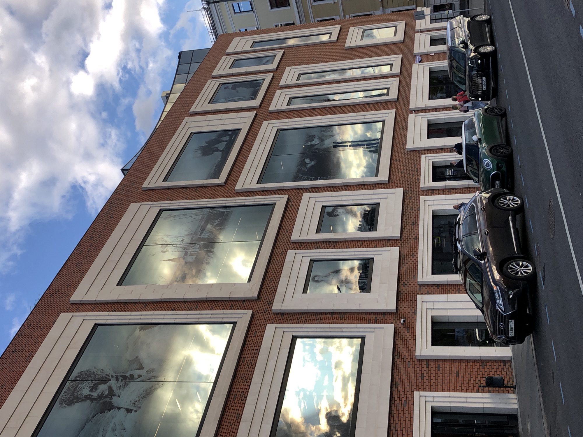 This is an exterior, eye-level, medium shot of a modern brick building in Moscow, Russia, with a cloudy sky in the background. The building features a grid of large, rectangular windows with light-colored frames. These windows display large black and white images, seemingly historical or artistic in nature, and also reflect the sky. Below the windows, on the ground level, are entrances to the building. In front of the building, parked along the curb of a street, are three cars: a dark brown Lexus SUV, a green Mini Cooper, and a black vehicle that appears to be a customized London black cab. Several pedestrians are visible near the entrances and interacting with each other. The street has painted lane markers.