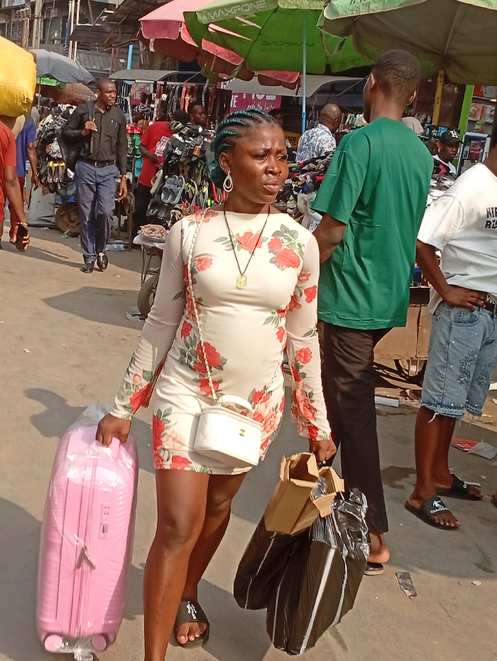 In a busy market scene, a woman stands out. She wears a floral dress, sandals, and a small white purse. With her hair braided, she carries a large pink suitcase in one hand and multiple bags in the other. She has a serious expression on her face, and stands among men in casual clothes. Vendors display their goods in the background.