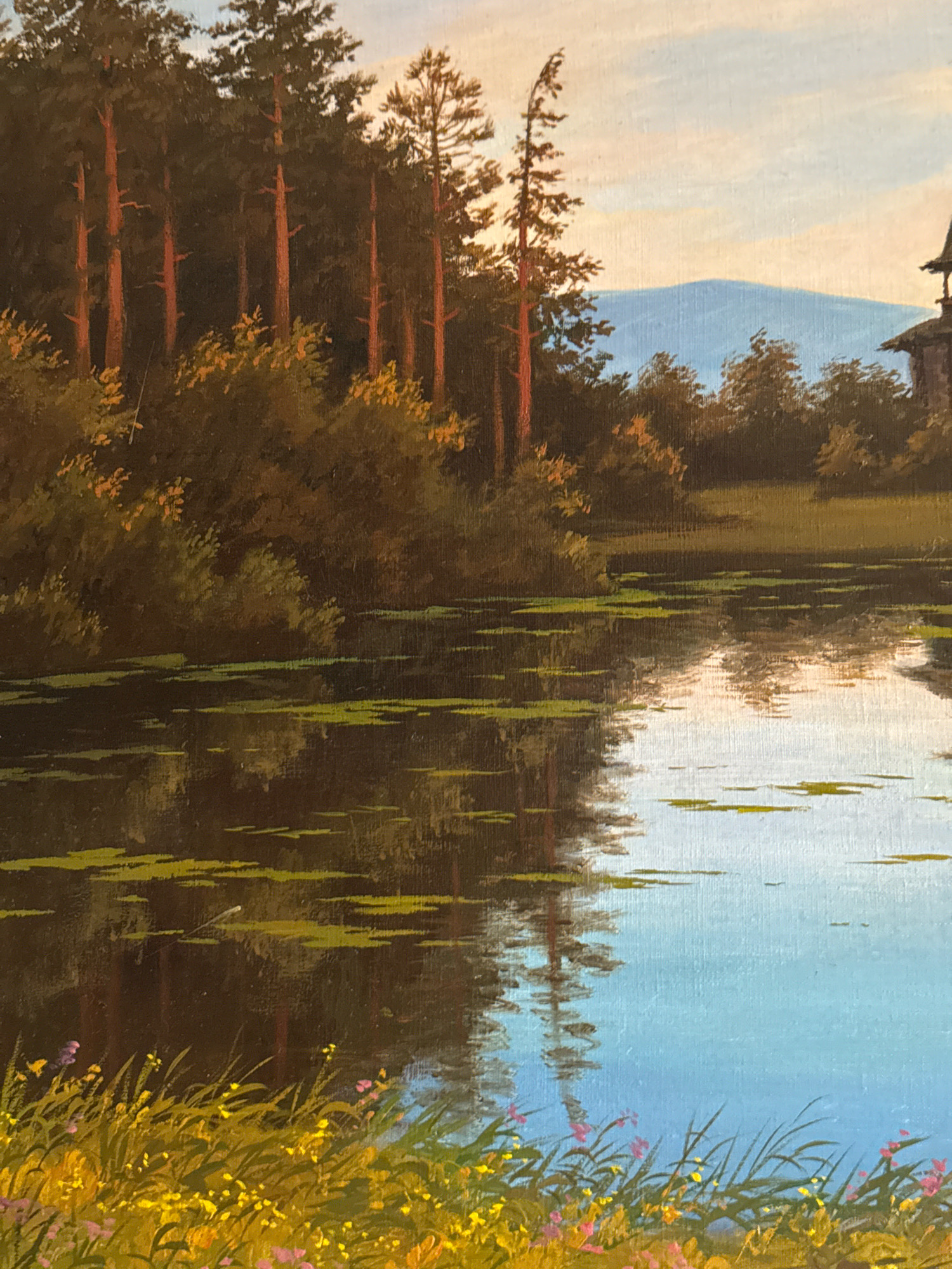 This is a painting depicting a natural landscape. In the foreground, there is a body of water, possibly a lake or pond, with lily pads or similar aquatic vegetation scattered across its surface. The water reflects the trees and sky. The middle ground features a dense stand of tall, slender trees with reddish-brown trunks, interspersed with foliage in shades of green and gold. Beyond the trees, there are rolling hills or mountains under a pale blue sky with wispy clouds. To the far right, a dark, wooden structure, possibly a building or part of a larger edifice with a pointed roof, is partially visible. The foreground also shows a patch of tall grass with small yellow and pink wildflowers. The painting is in a realistic style. There are no people or animals visible in the scene. The image does not provide any specific information about the city of Krasnoyarsk, Russia.