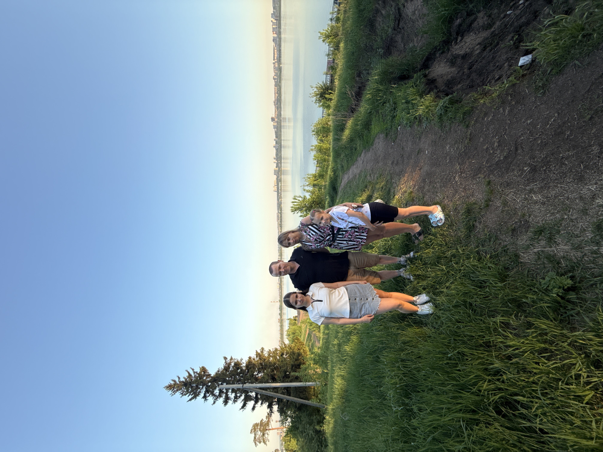 A group of four people is standing on a grassy hill overlooking a body of water, which appears to be a river or lake, with a city skyline in the background. The city is Izhevsk, Russia. From the top to bottom:
- An adult woman with light-colored hair is wearing a knee-length dress with a pattern of black and white.
- A young girl is wearing a white t-shirt with a floral print, black shorts, and light-colored sandals. The woman is embracing her from the back.
- A middle-aged man with short dark hair is wearing a black t-shirt and khaki shorts.
- A young woman is wearing a white collared shirt, a gray skirt, and white sneakers.
The environment consists of a grassy hillside with a dirt path, a tree to the lower-left side. The sky is clear and blue.